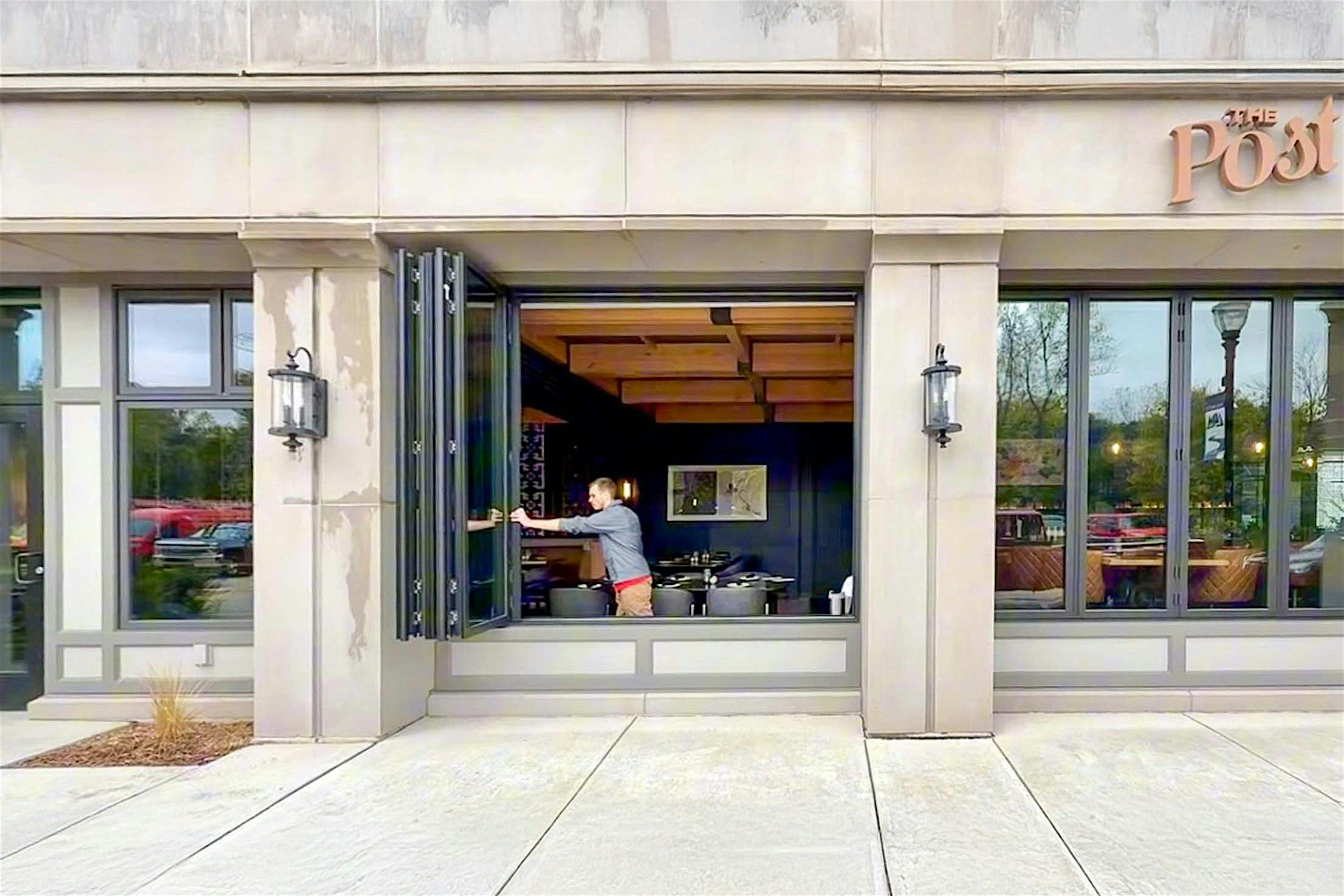 A person swings open sleek commercial bifold windows at the front of the Post Tavern, revealing the inviting interior with neatly arranged tables and chairs.