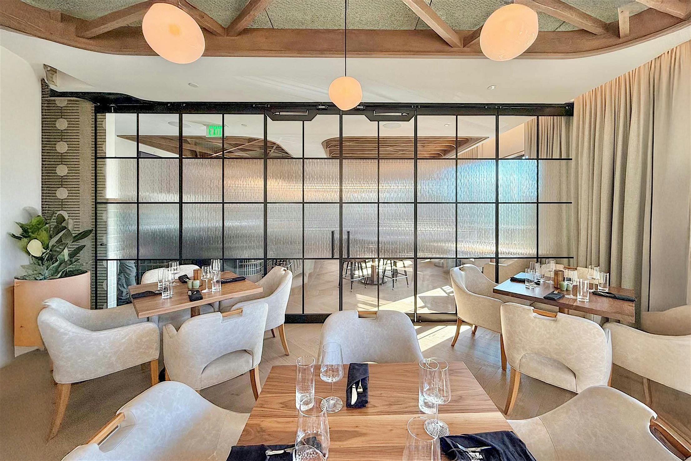 A modern restaurant interior features light wood tables and beige chairs, enhanced by a large glass wall partition for a future amenities area.