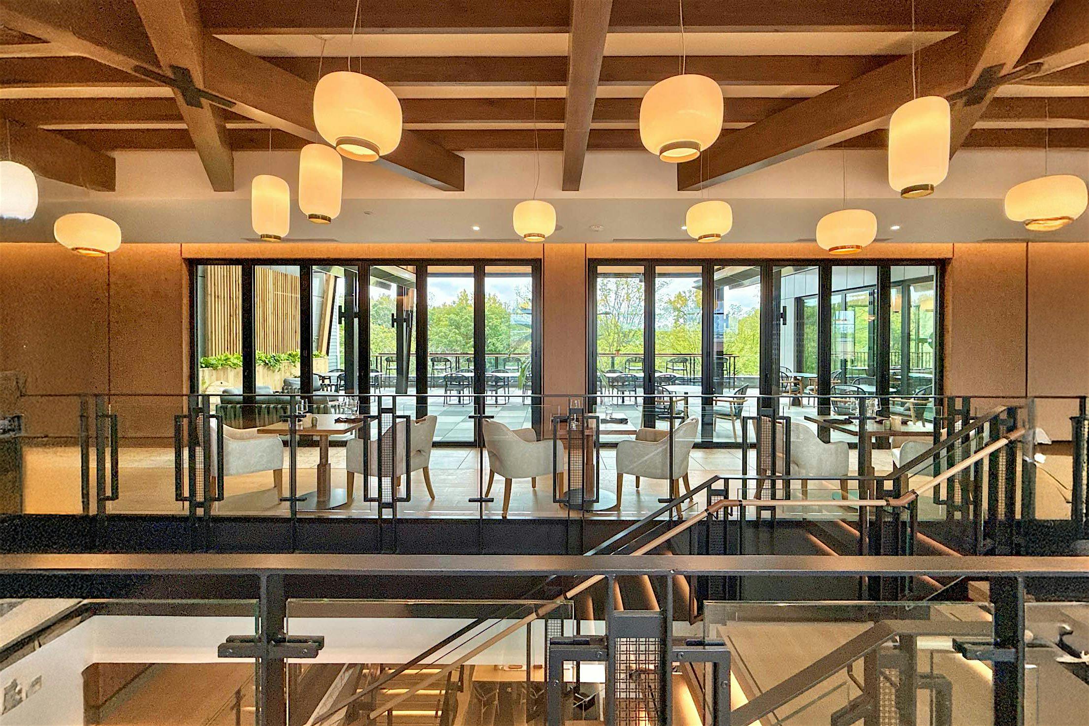 Modern restaurant interior with wooden beams and hanging round lights, featuring tables and chairs set by large bifold glass doors.