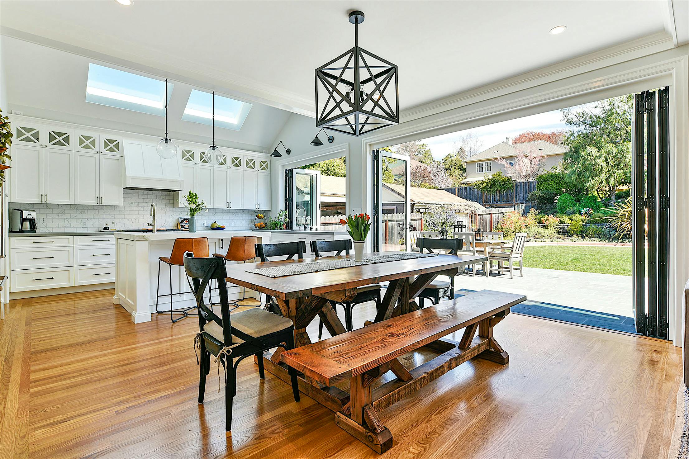 Contemporary open-plan kitchen and dining area with wooden floors, modern white cabinets, skylights, and folding glass walls leading to a backyard patio.