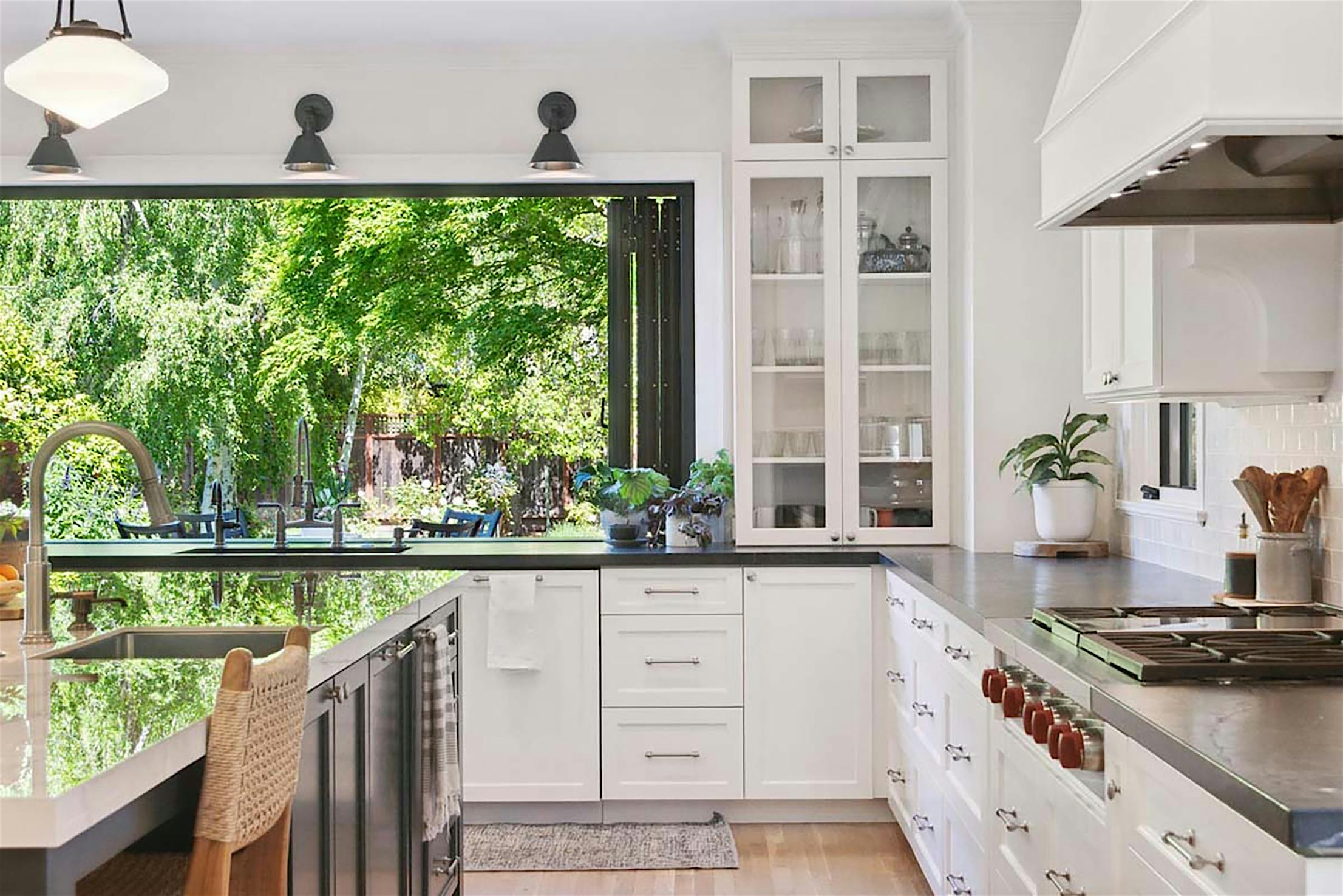 Modern kitchen with white cabinets, black countertops, and a large folding glass door opens to a garden view, seamlessly blending indoor and outdoor spaces.