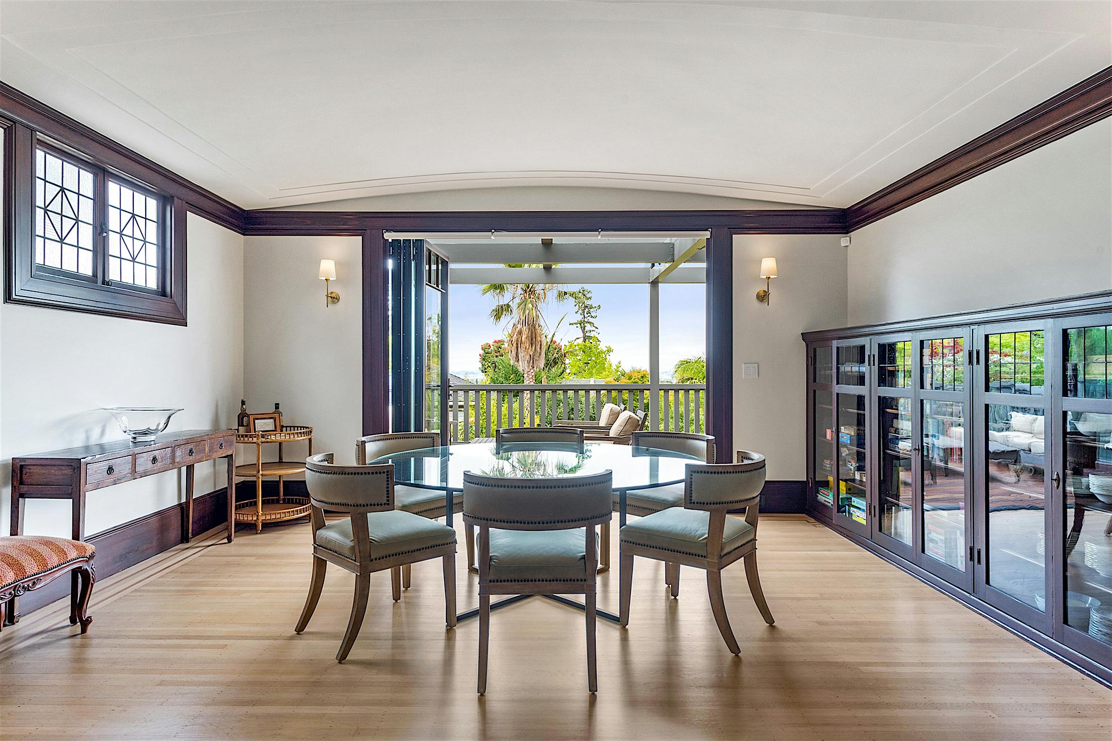 A dining room with a round table and six chairs features sleek folding glass doors that open to a terrace with two outdoor chairs, seamlessly blending the indoor space with the serene outdoors.