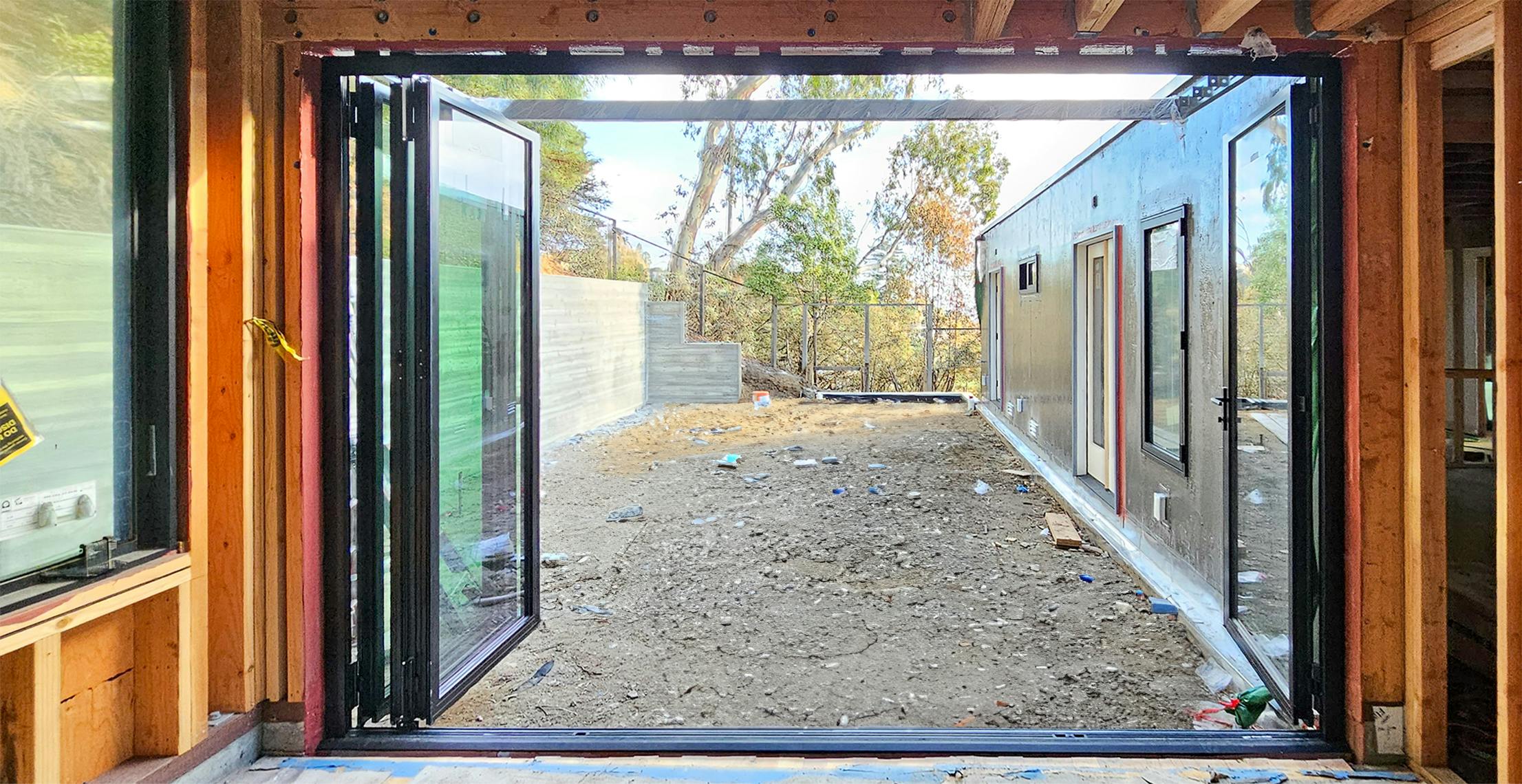 Open aluminum clad folding glass wall reveals a yard under construction, framed by wooden walls and trees in the background.