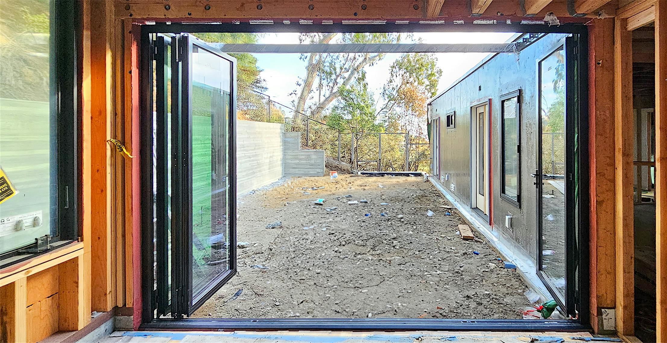 Open aluminum clad folding glass wall reveals a yard under construction, framed by wooden walls and trees in the background.