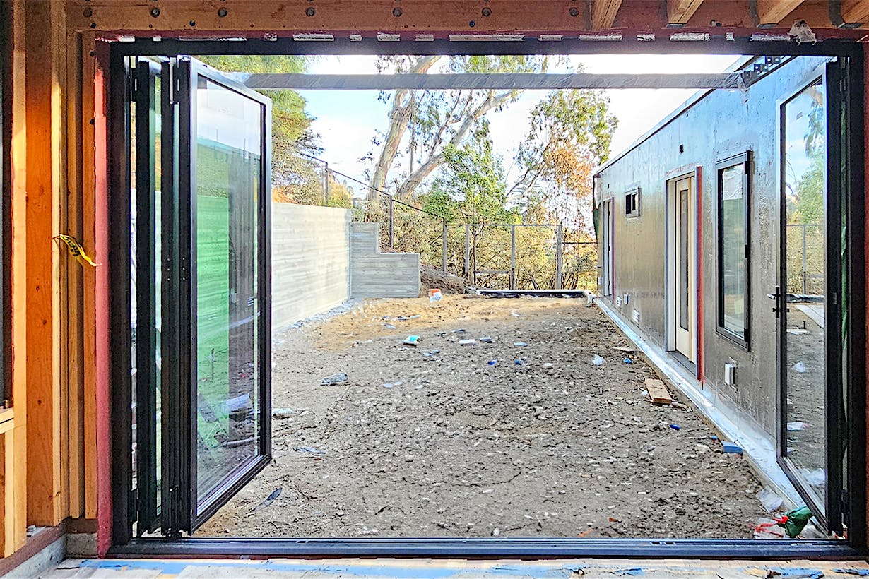 Open aluminum clad folding glass wall reveals a yard under construction, framed by wooden walls and trees in the background.