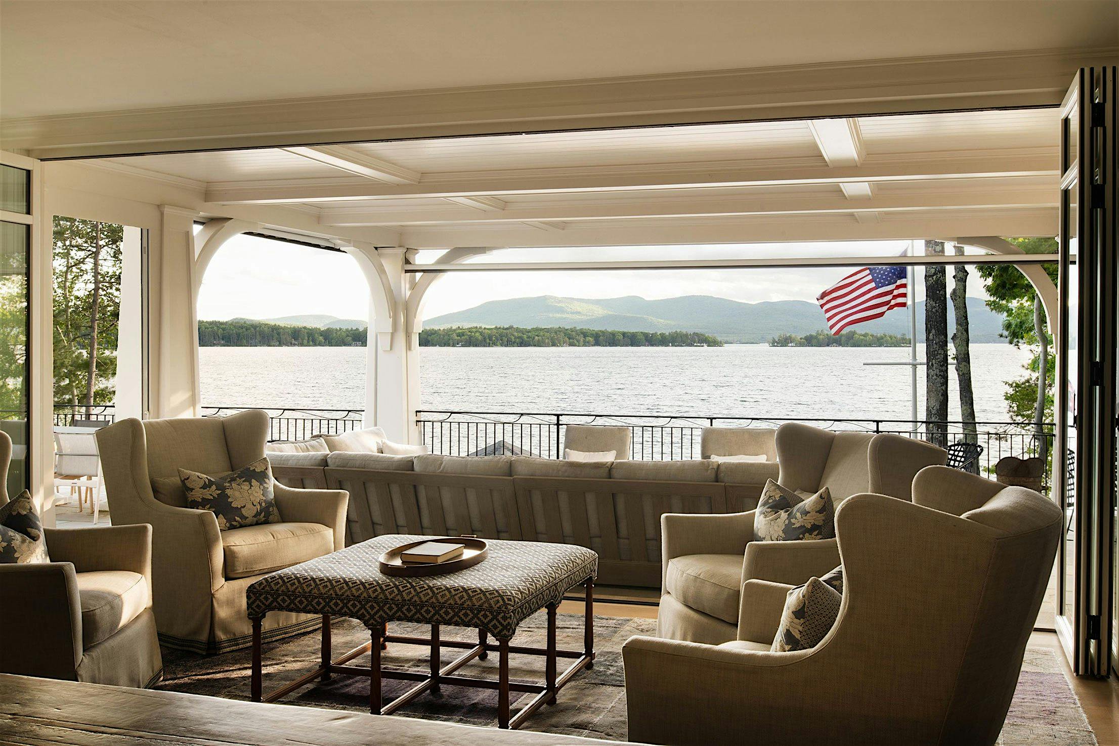 Living room with beige armchairs and sofa facing large patio glass doors, offering a breathtaking view of a lake and mountains.