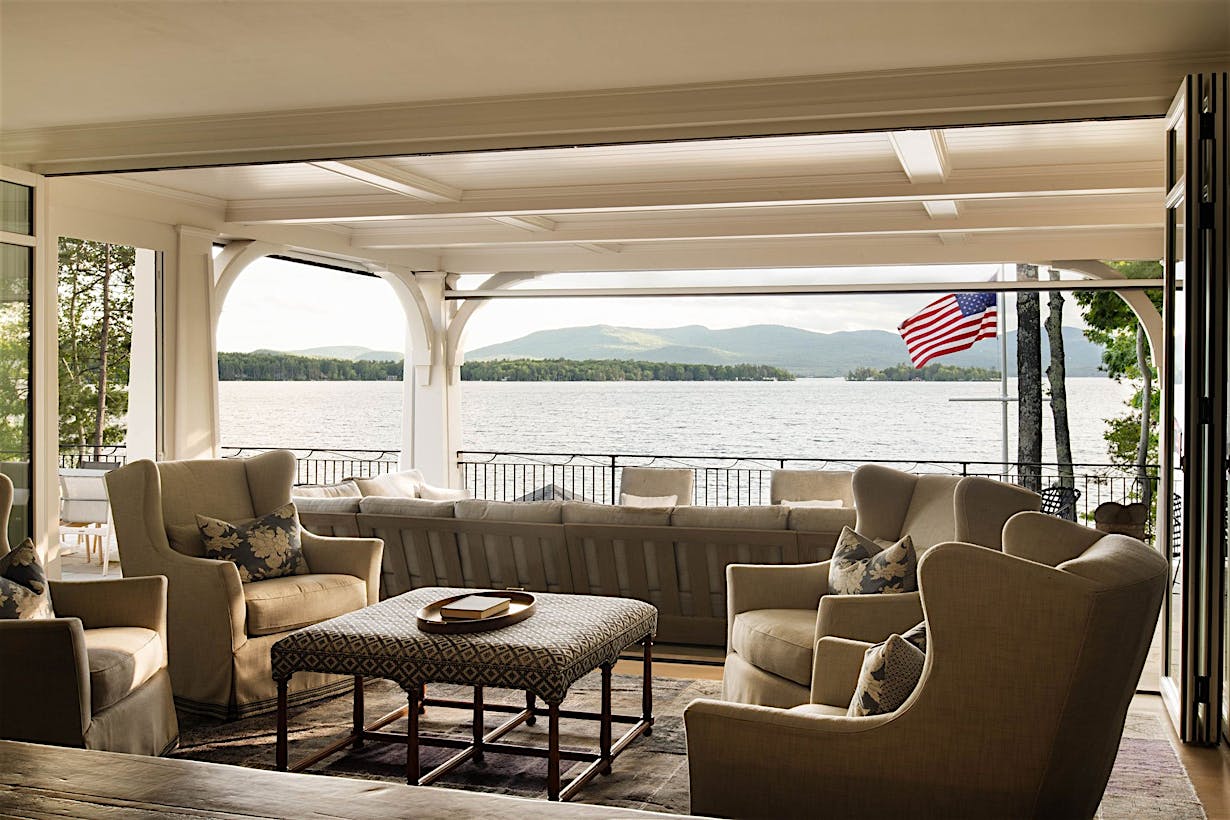 Living room with beige armchairs and sofa facing large patio glass doors, offering a breathtaking view of a lake and mountains.