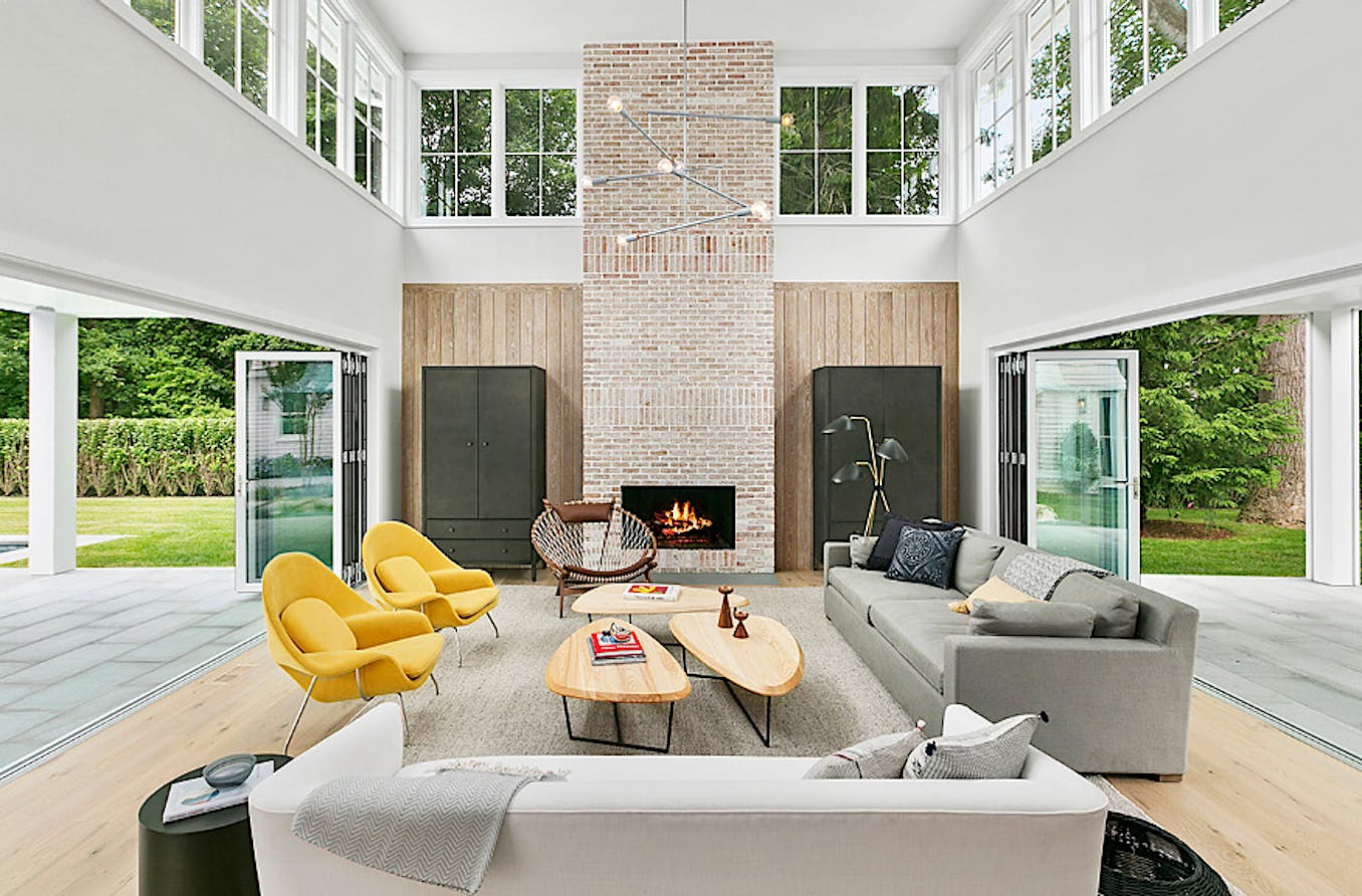 A modern living room with high ceilings features a brick fireplace, a gray sofa, and two large folding glass doors opening to a lush outdoor space.