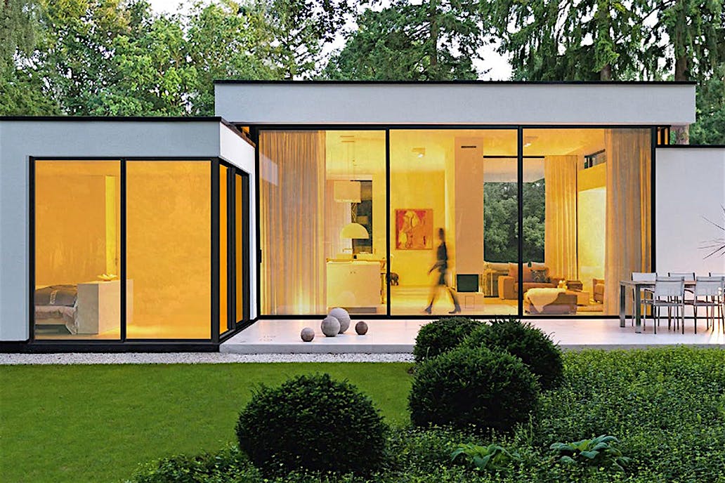 A modern house features large glass windows and a sleek cero minimal sliding glass door system, glowing warmly from indoor lights.