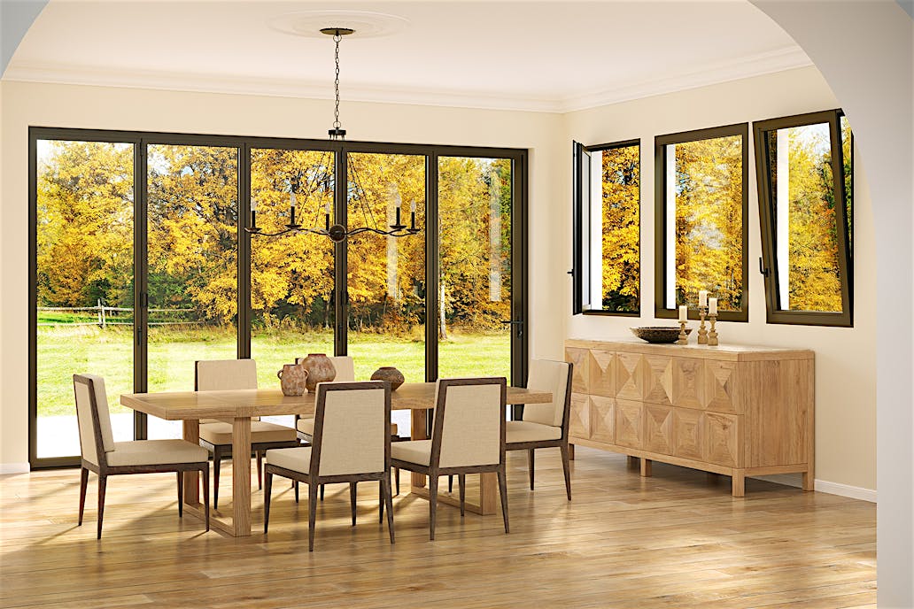 A dining room with a wooden table, six chairs, and a sideboard features Generation 4 folding and sliding glass walls and tilt turn windows, offering a uniform aesthetic and framing views of trees with yellow autumn leaves outside.
