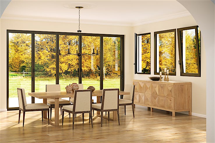 A dining room with a wooden table, six chairs, and a sideboard features Generation 4 folding and sliding glass walls and tilt turn windows, offering a uniform aesthetic and framing views of trees with yellow autumn leaves outside.