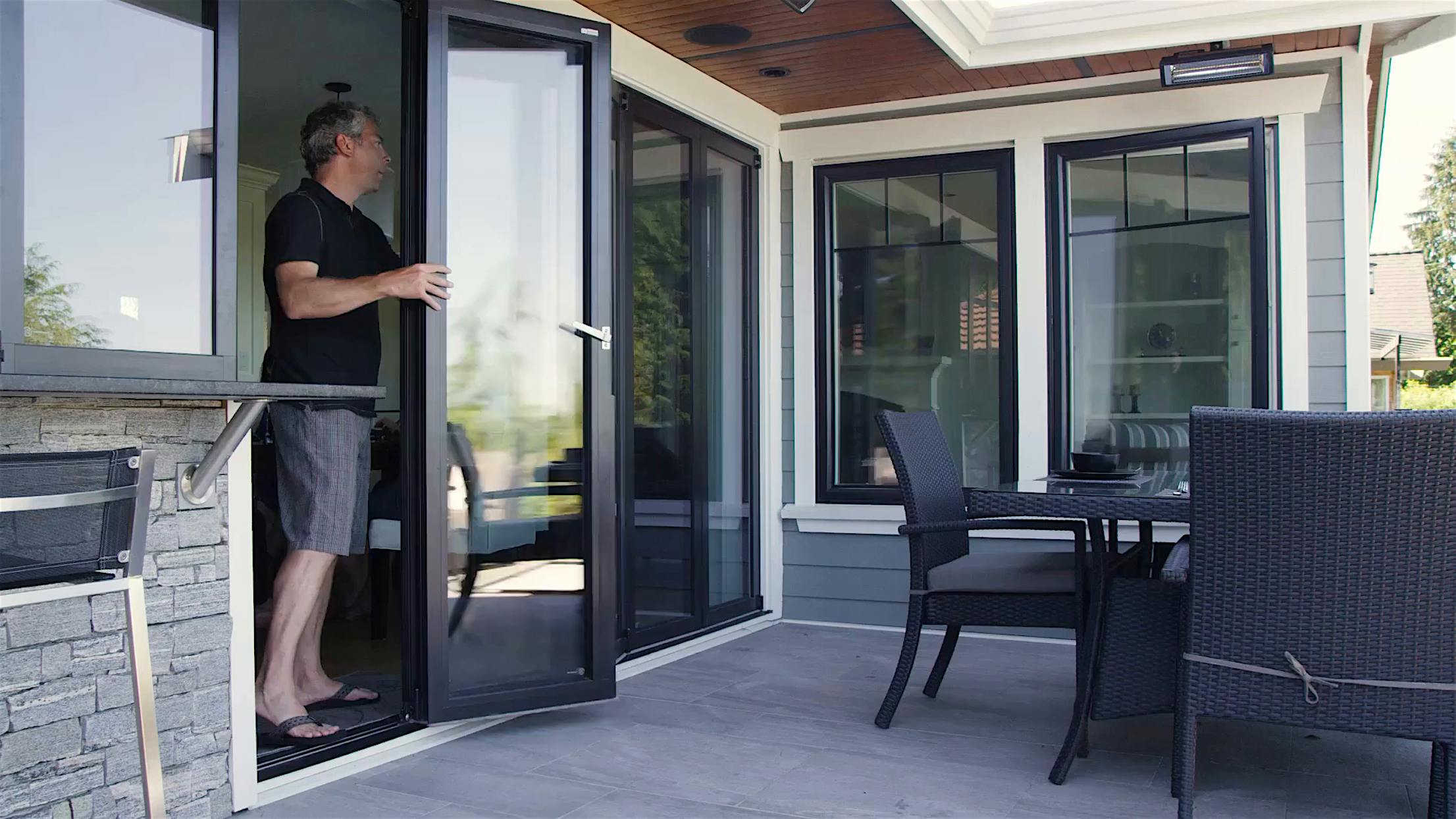 Modern indoor-outdoor kitchen design. A man effortlessly glides open sleek folding glass doors, revealing a stunning outdoor patio complete with stylish seating and a welcoming dining area.