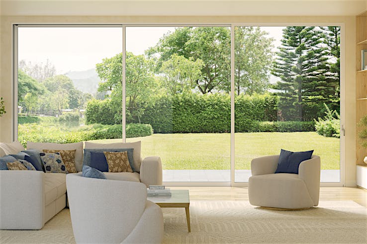A modern living room with white sofas and a wooden coffee table, featuring slim aluminum framed multislide glass doors overlooking a green garden with trees and shrubs.
