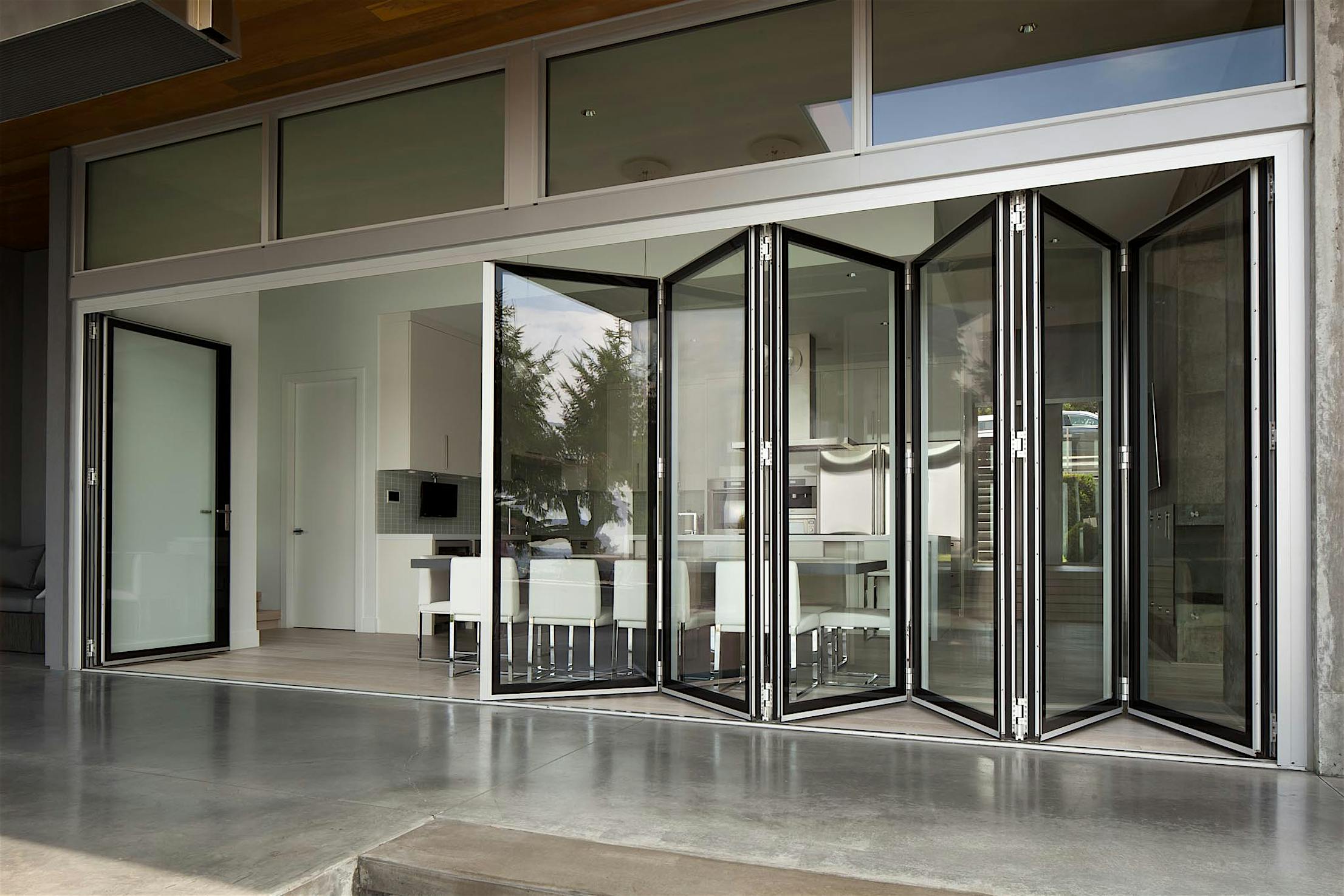Glass folding doors open to reveal a modern kitchen and dining area. White cabinets and a table with chairs are visible through the doors.