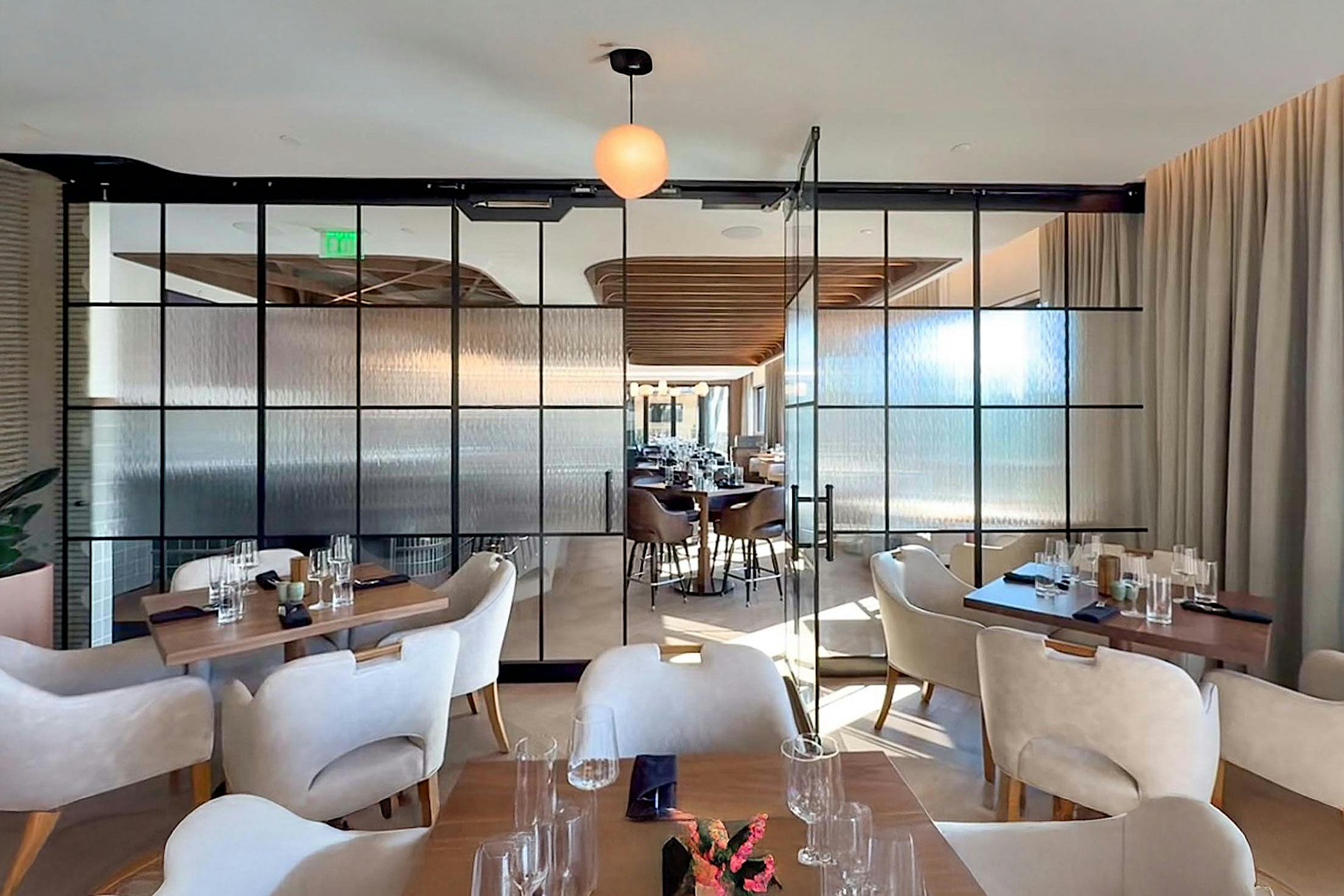 Modern restaurant with light-colored chairs, wooden tables, sliding glass partitions, and set dining tables, seen through partially open swing doors.