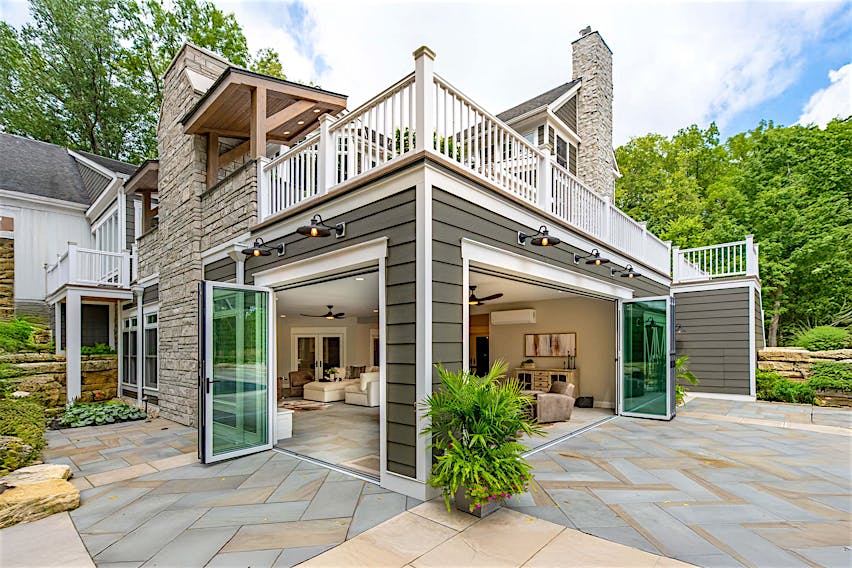 Modern house with stone and siding exterior, bifold glass doors open to a patio, upper balcony with white railing, and surrounding greenery.