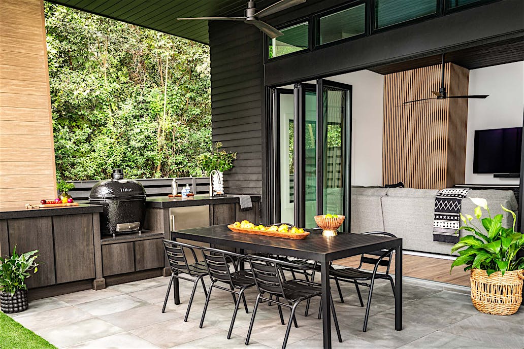 Modern outdoor patio with black dining table and six chairs, built-in grill, countertop, and and large folding glass doors opening to a living room with a sofa and TV.