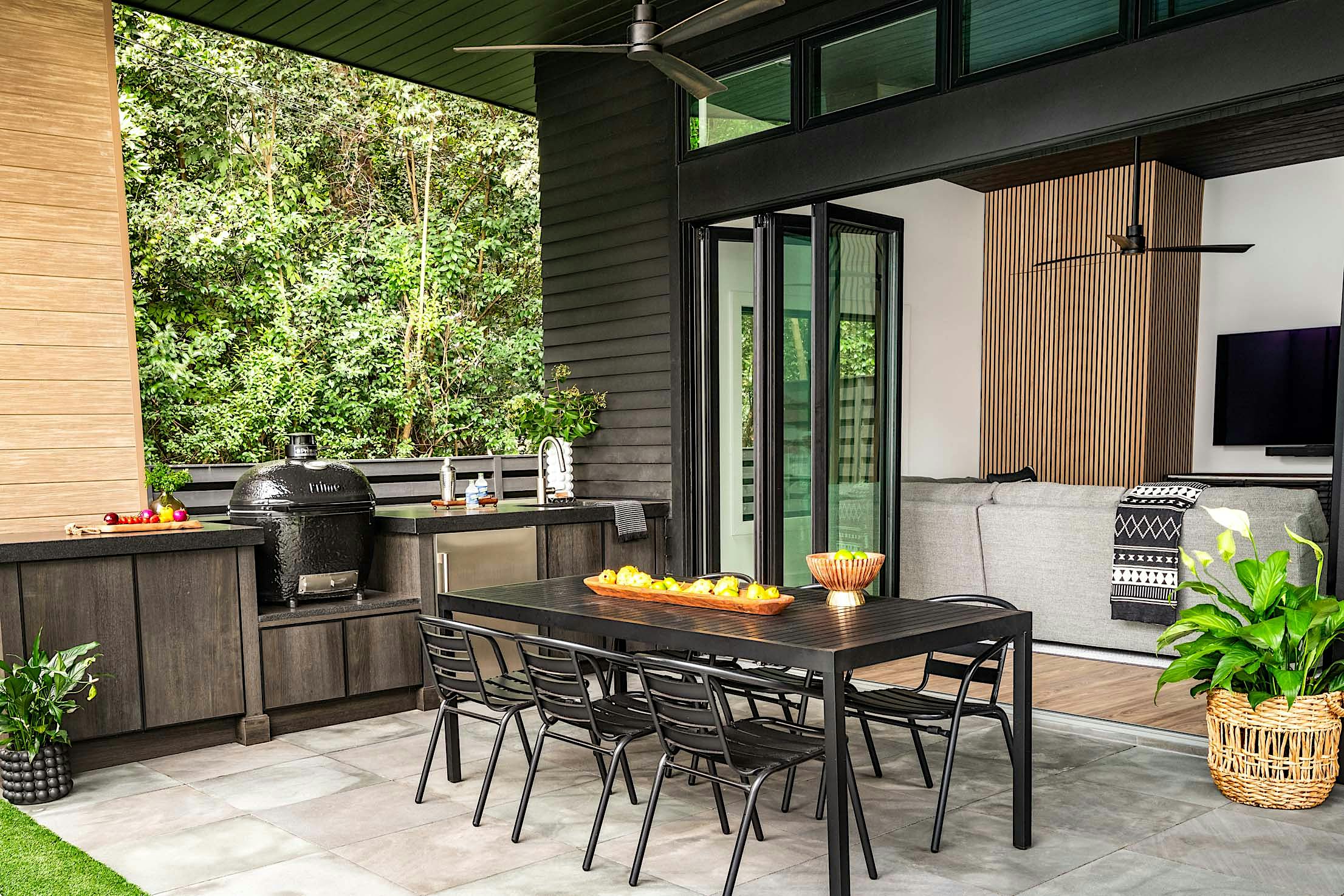 Modern outdoor patio with black dining table and six chairs, built-in grill, countertop, and and large folding glass doors opening to a living room with a sofa and TV.
