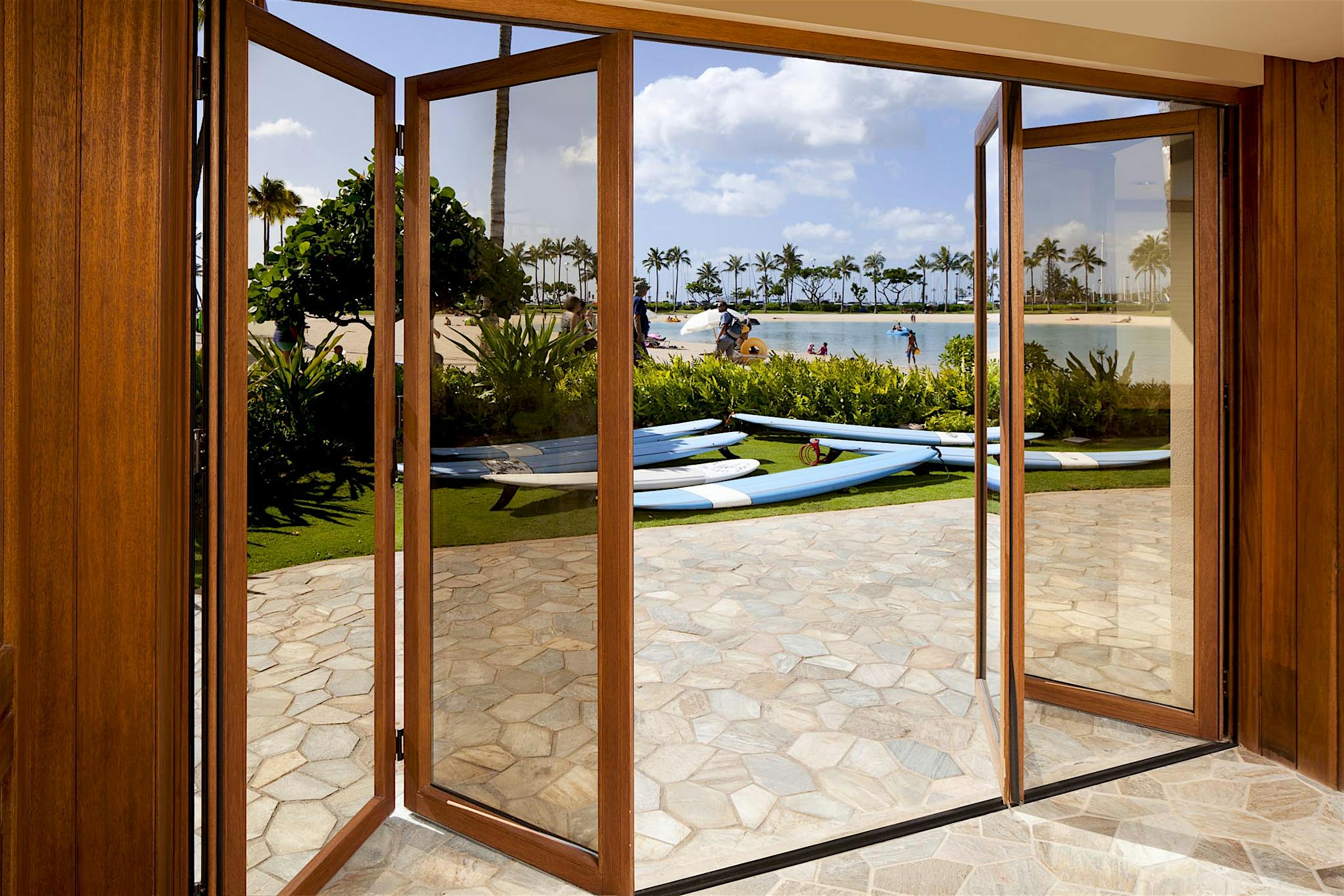 Open glass bifold doors lead to a patio with a view of a beach. Paddleboards rest on the grass, and people are lounging near the water under a partly cloudy sky.
