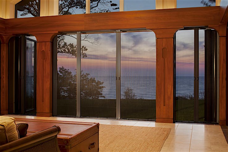 Large glass doors with fully retractable screen panels offer a view of a sunset over a body of water, framed by wooden trim inside a living room.