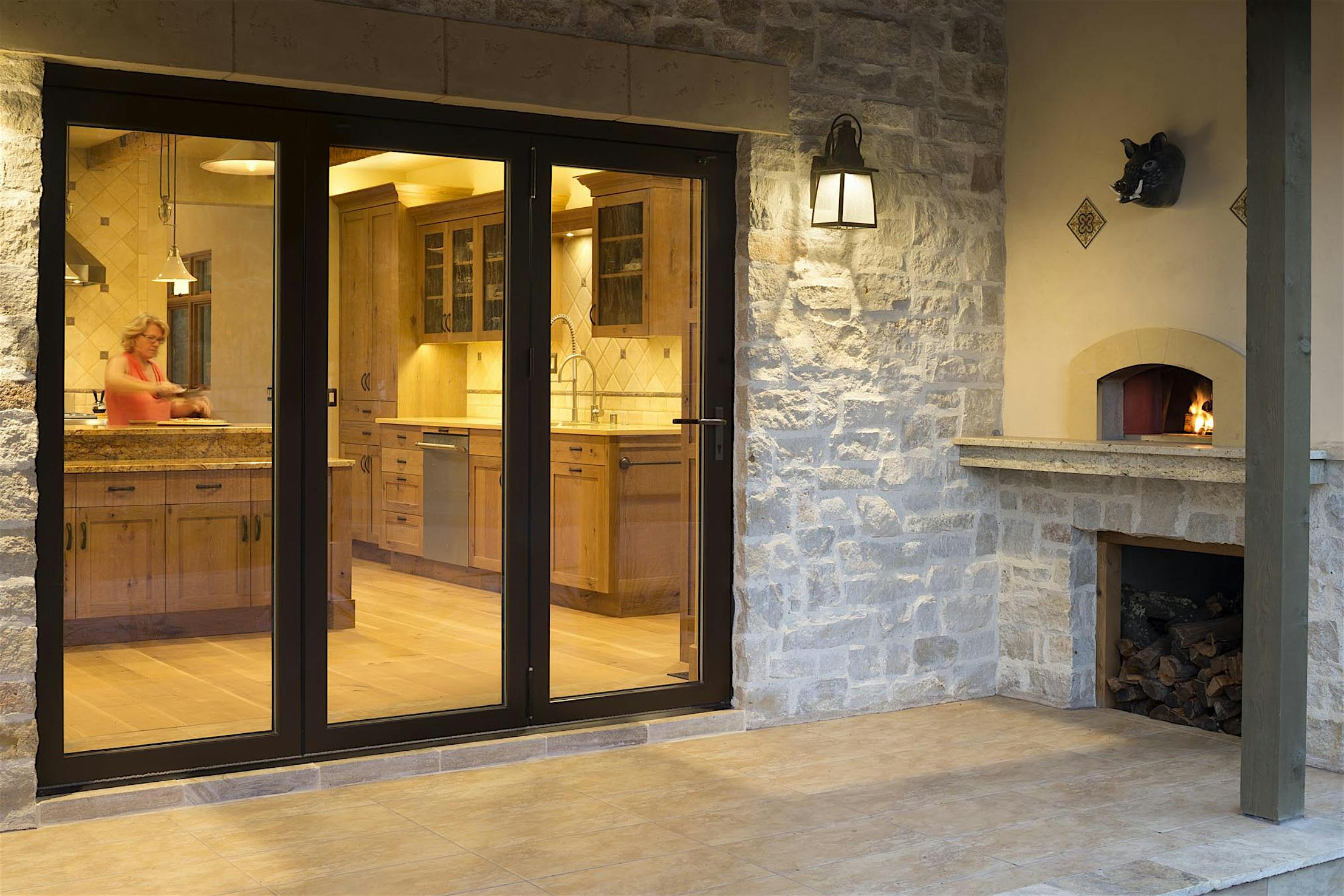 A person is working in a kitchen viewed through large folding glass doors. Outside, there is a stone fireplace with a small fire burning, seamlessly connecting the cozy indoor space with the inviting outdoor ambiance.