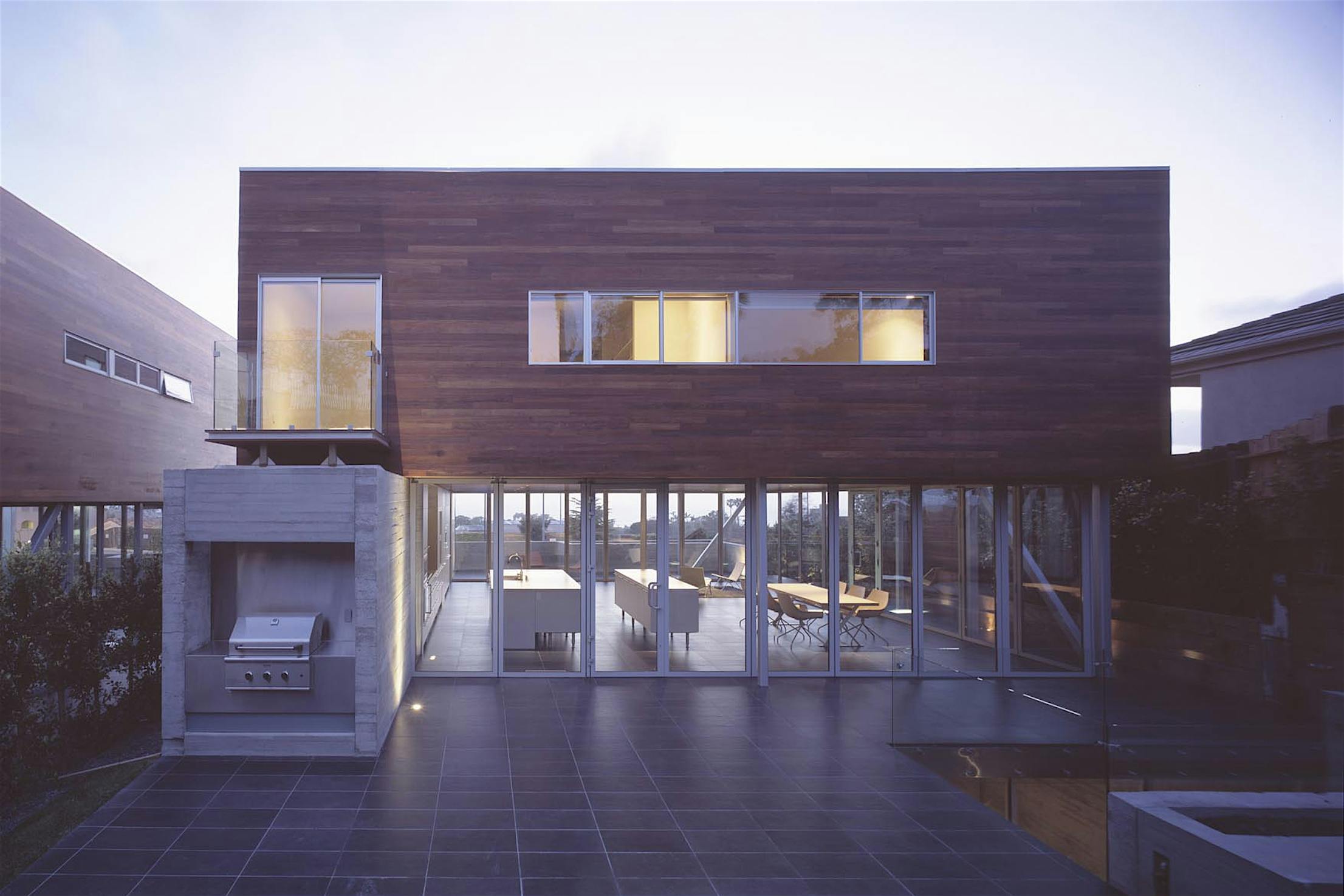 Modern two-story house with large windows, a wooden exterior accent, and an outdoor patio featuring a built-in grill and sliding glass patio doors.