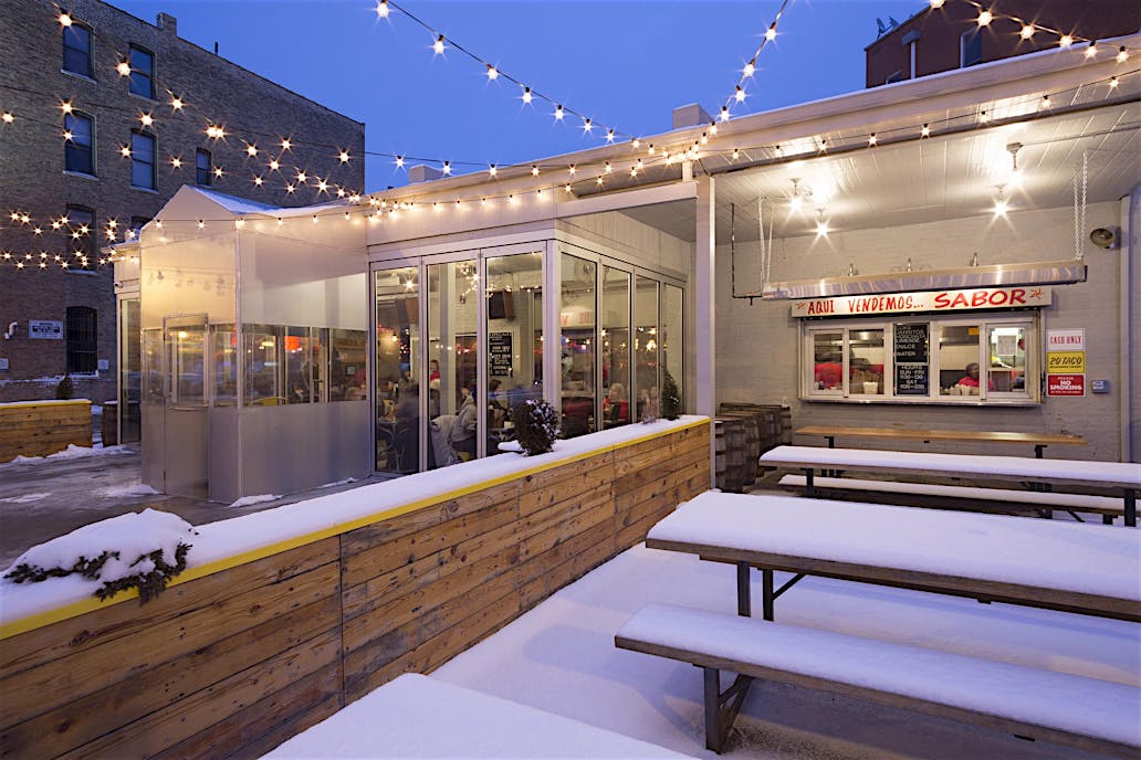 Outdoor restaurant patio with string lights, snow-covered seating, and high-performance glass doors showcasing a cozy indoor restaurant space.