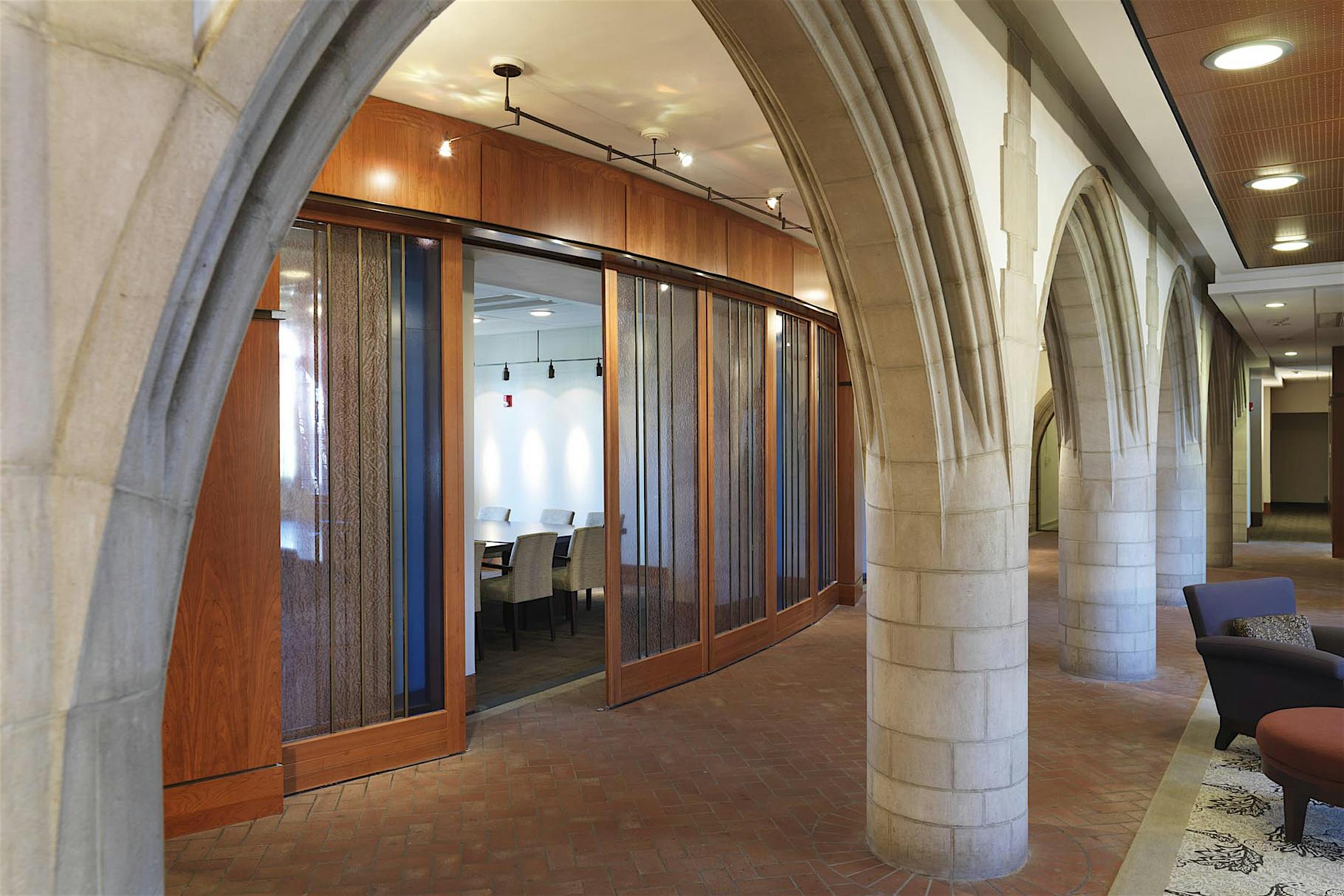 Stone arches line a brick-floored hallway leading to a glass-walled conference room with partially open wood-framed glass doors and white chairs.