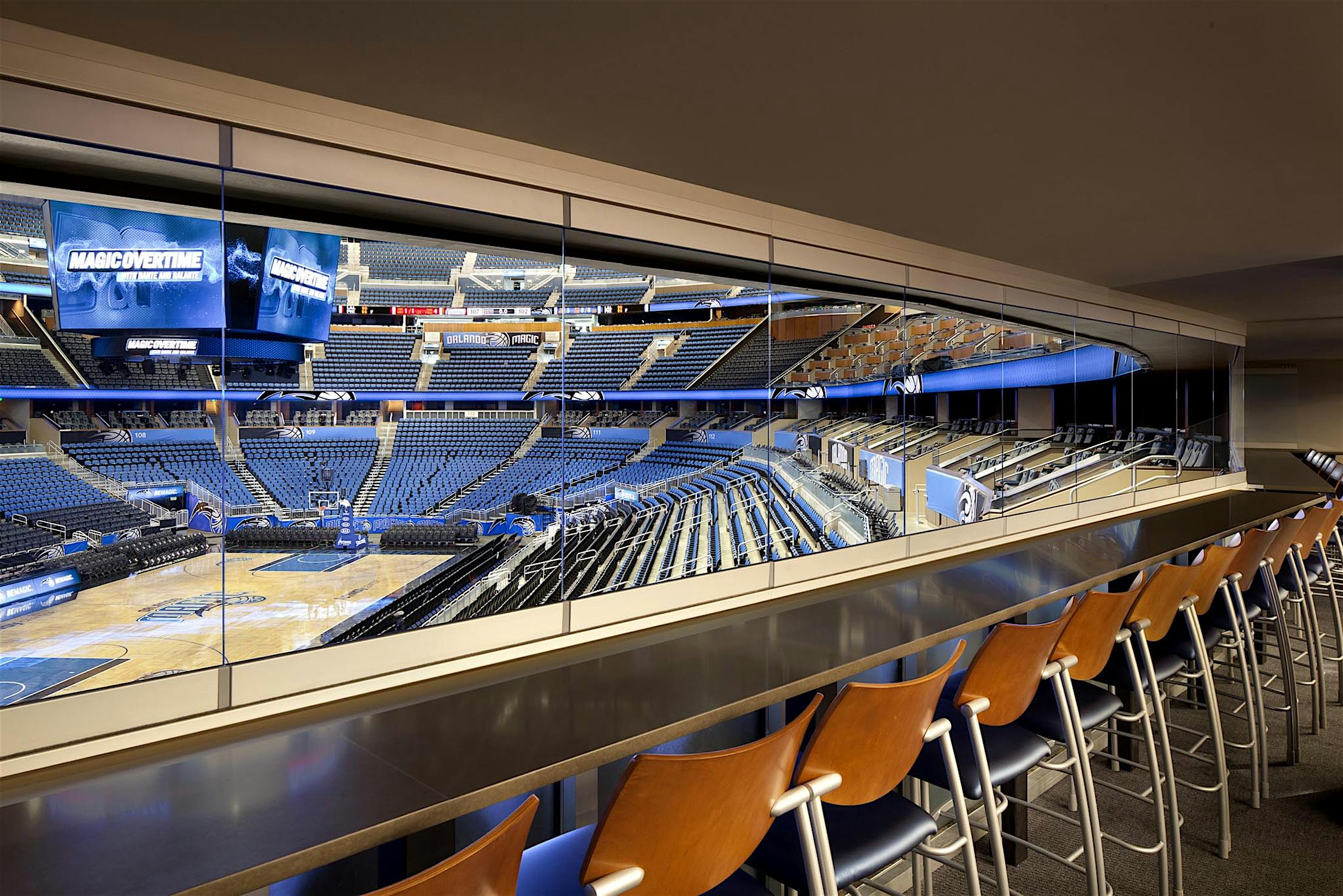Orlando Magic suite with seating and frameless sliding glass windows, overlooking the indoor basketball arena and scoreboard.