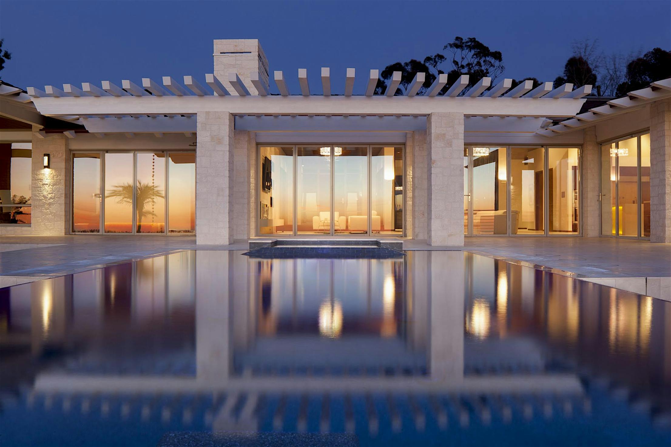 Modern house with large bifold glass doors and a pergola-covered patio overlooking a pool at dusk.