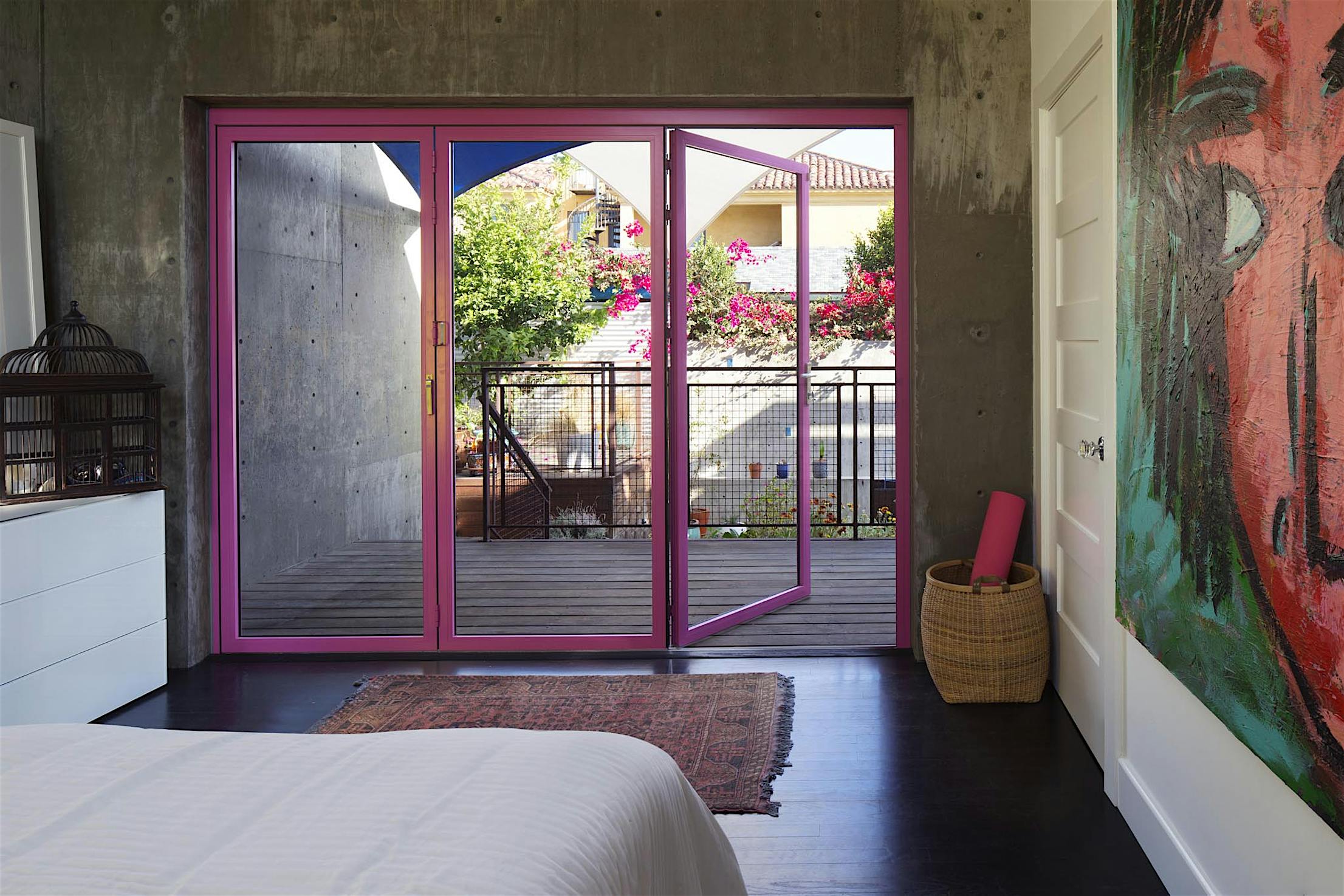 Modern bedroom with concrete accent wall, custom-colored glass swing doors opening to a balcony, and a vibrant abstract painting on the side wall.