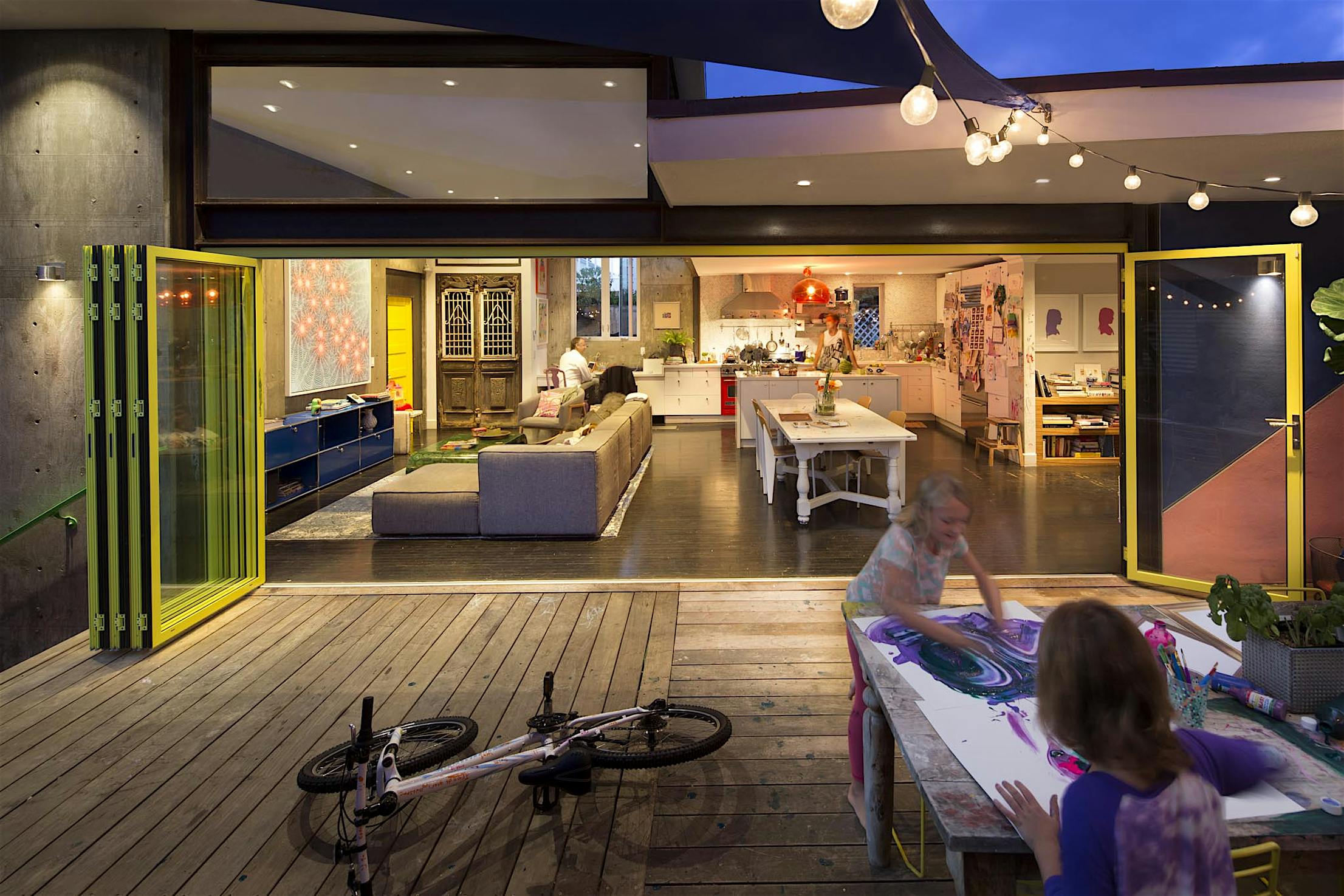 Two children make art on a wooden deck near accordion glass doors opening to a modern, well-lit living and kitchen area.