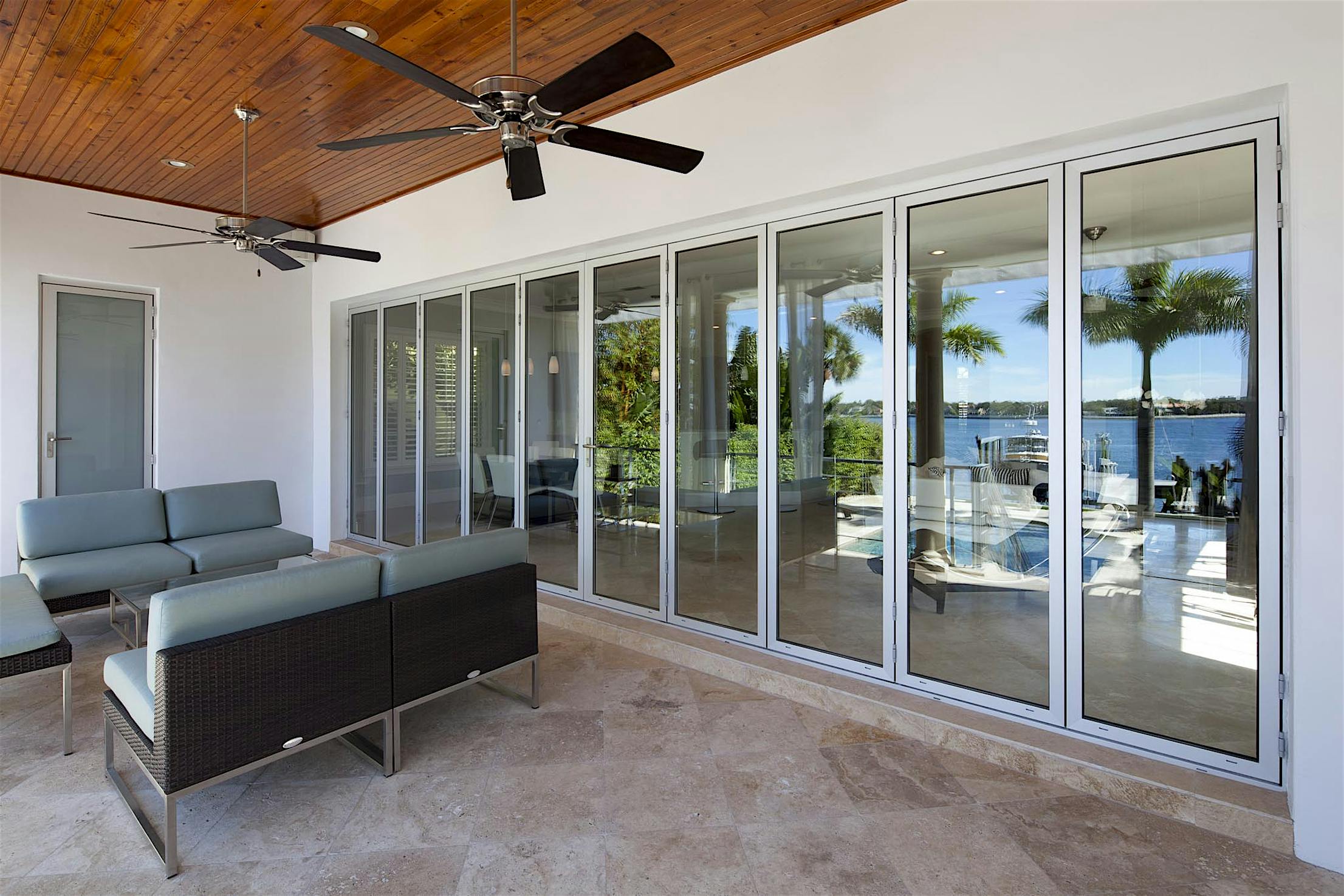 Modern patio seating area with aluminum-framed folding glass walls offering a view of a lake and palm trees.