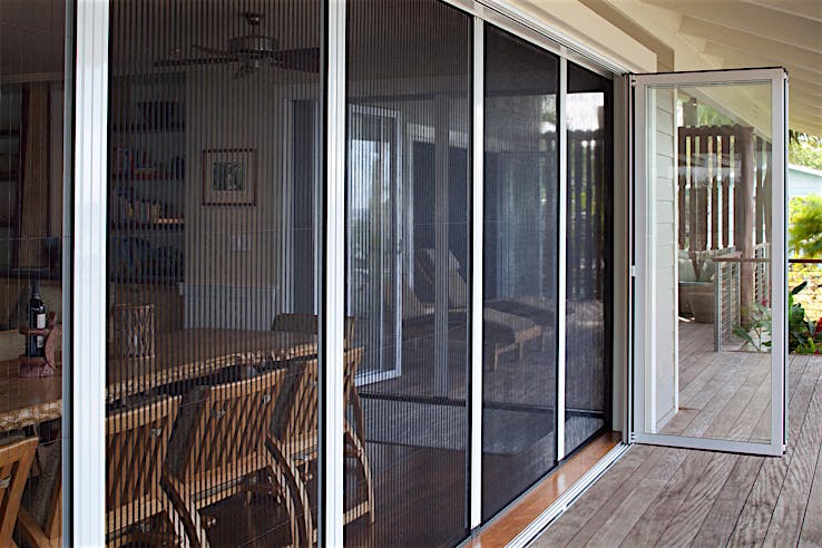 A patio with bifold glass doors and a fully retractable screen door leads from a wooden deck into a dining area with wooden chairs and a table.