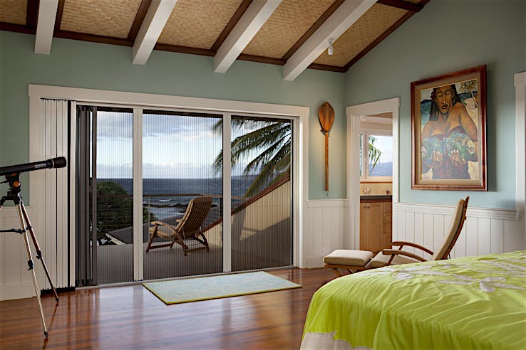 Bedroom with wooden floors, a green bedspread, and folding glass doors with a fully retractable screen opening to a balcony with a lounge chair and ocean view.