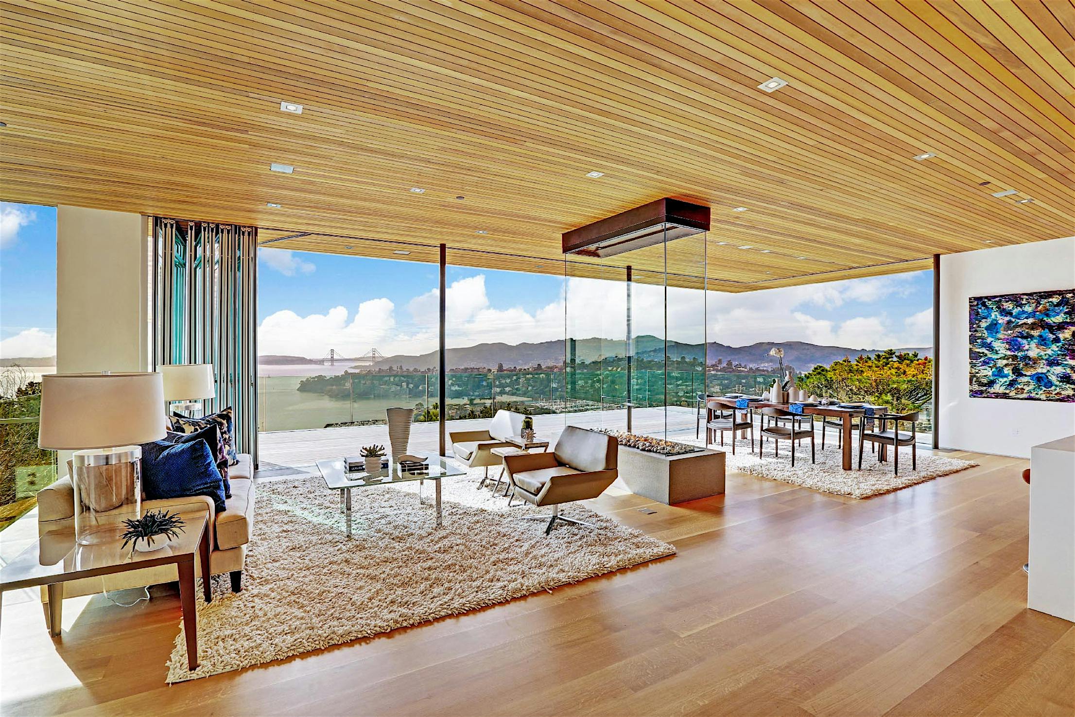 A modern living and dining area with floor-to-ceiling windows offering a panoramic view of a bay, bridge, and distant cityscape.