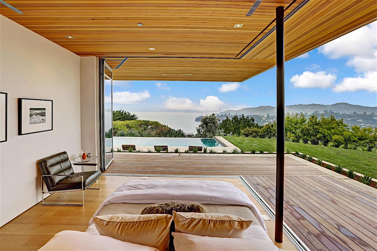Modern bedroom with corner sliding glass doors leading to a deck, lawn, infinity pool, and views of hills.
