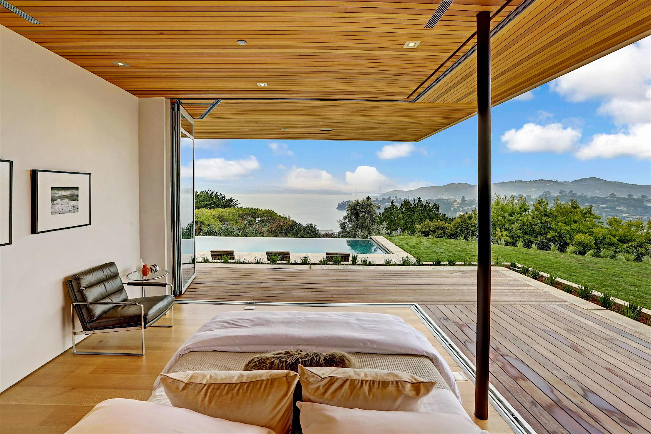 Modern bedroom with corner sliding glass doors leading to a deck, lawn, infinity pool, and views of hills.