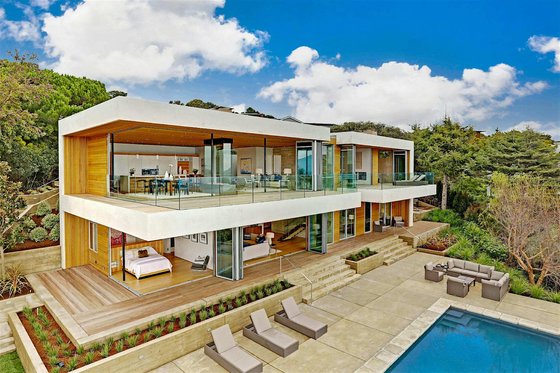 Modern two-story house with large sliding glass doors, open balconies, poolside loungers, and outdoor seating in landscaped surroundings.