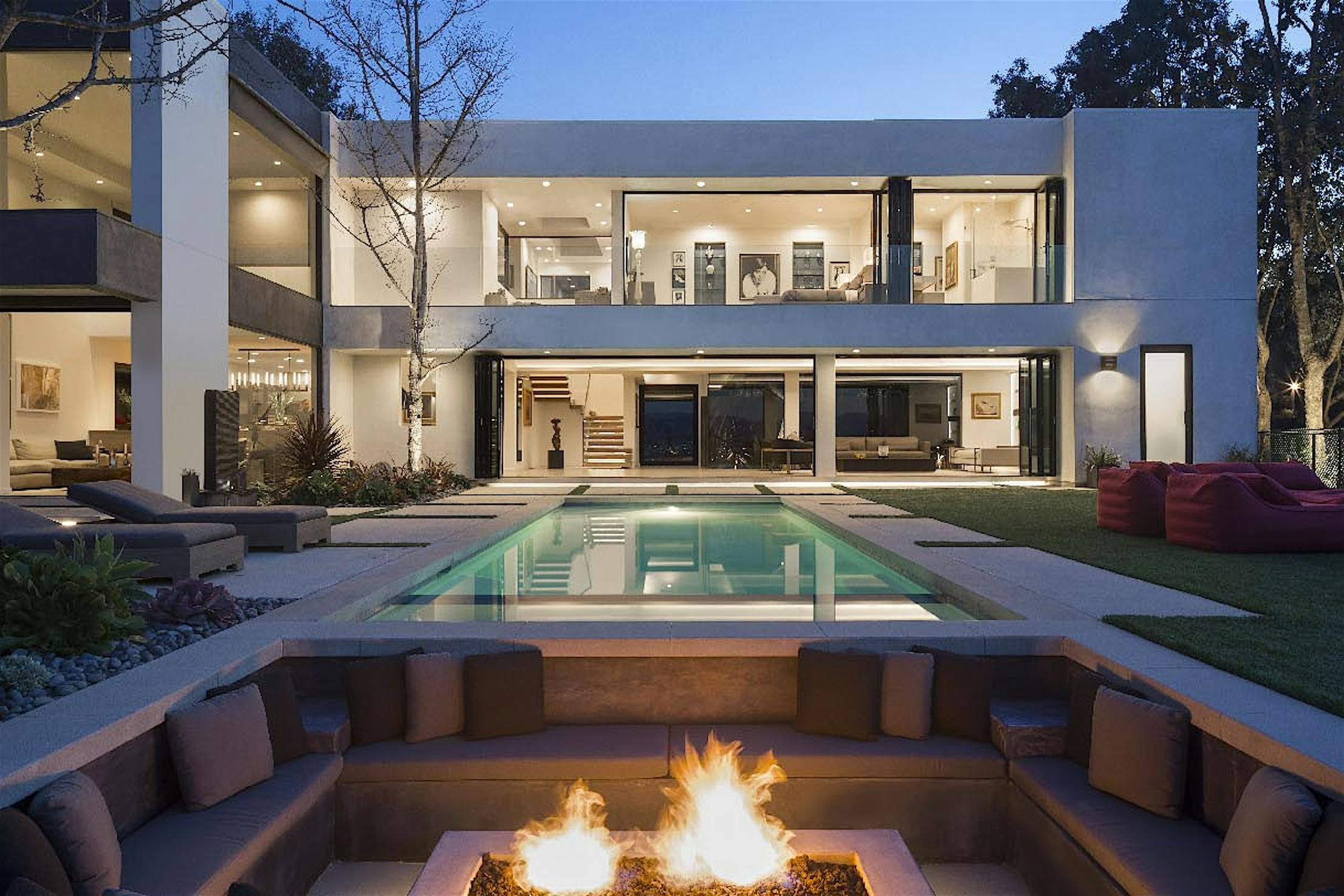 Modern two-story house with large glass windows and an elegant glass exterior doors, featuring a central swimming pool, surrounding seating areas, and a fire pit in the foreground, all illuminated at twilight.