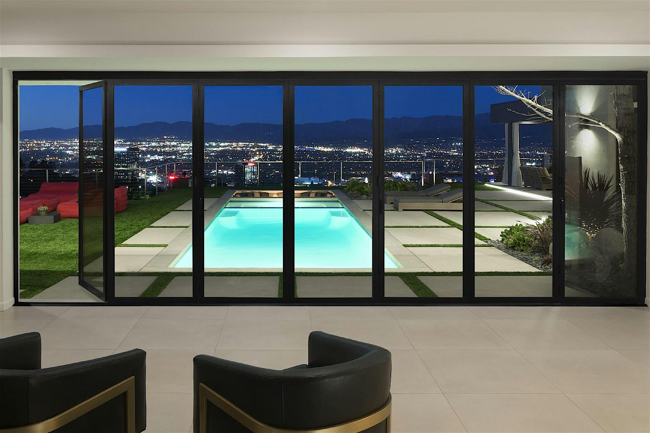 Modern living room interior with swing glass doors opening to a luxury pool, stylish patio, and panoramic nighttime cityscape view with mountain backdrop.