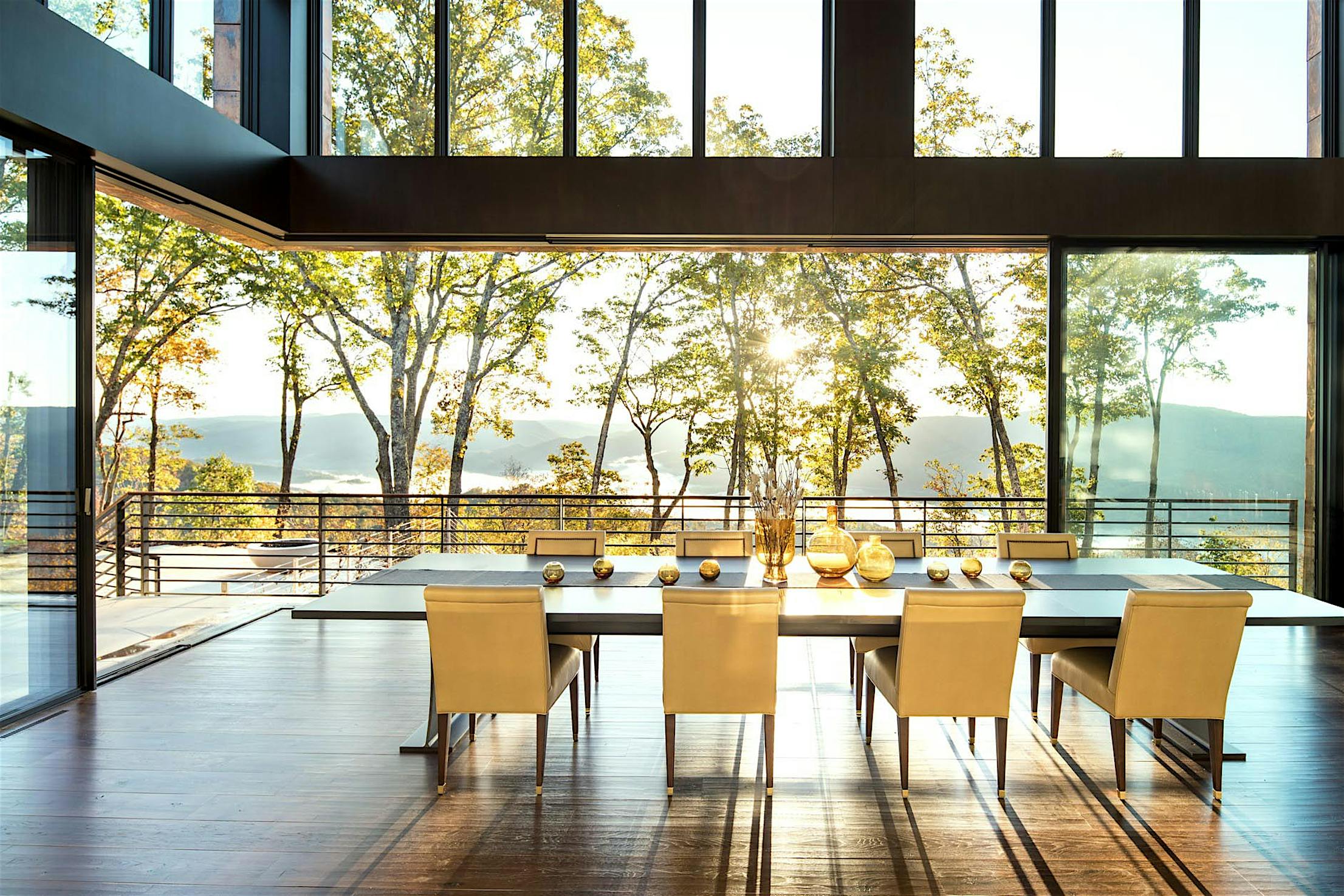 A modern dining room with a large table and eight chairs sits near large panel minimal sliding glass walls, offering views of trees and hills.