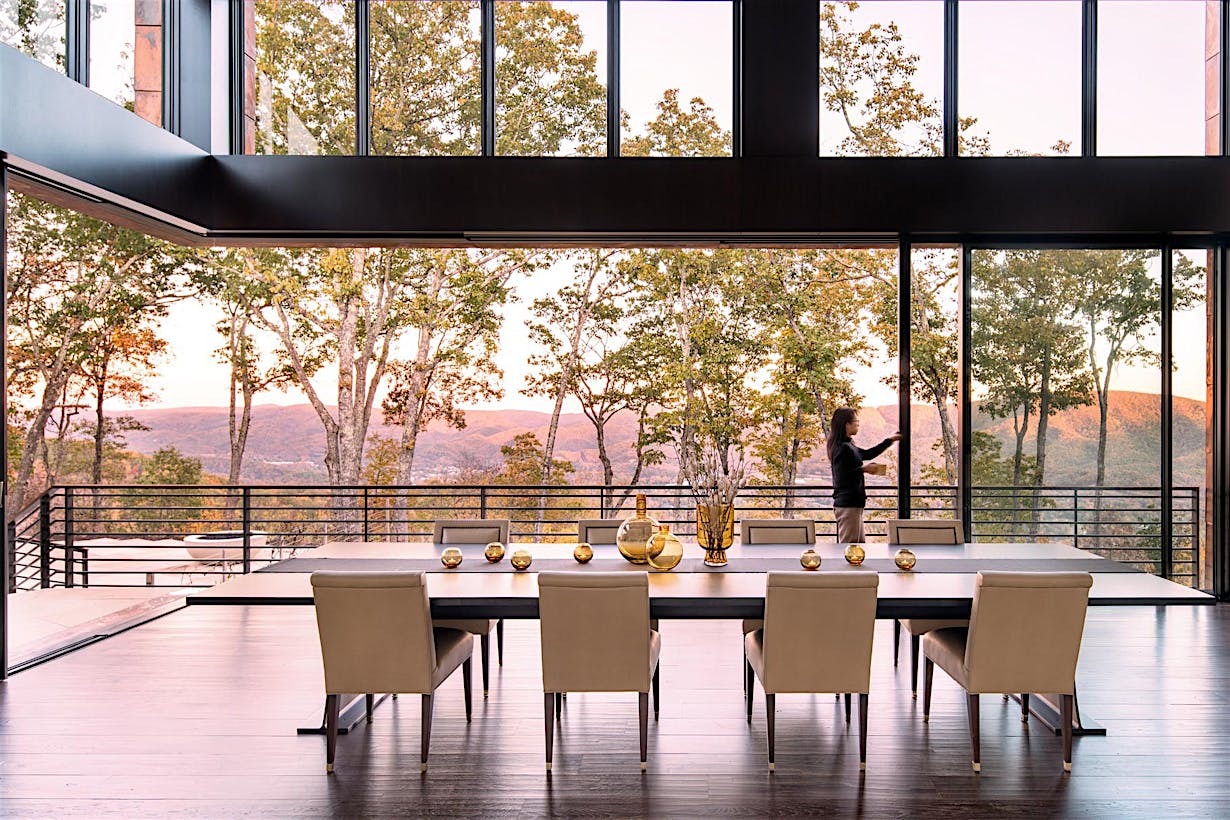A modern dining room with a large table and eight chairs sits near large panel minimal sliding glass walls, offering views of trees and hills; a person stands outside on the balcony.