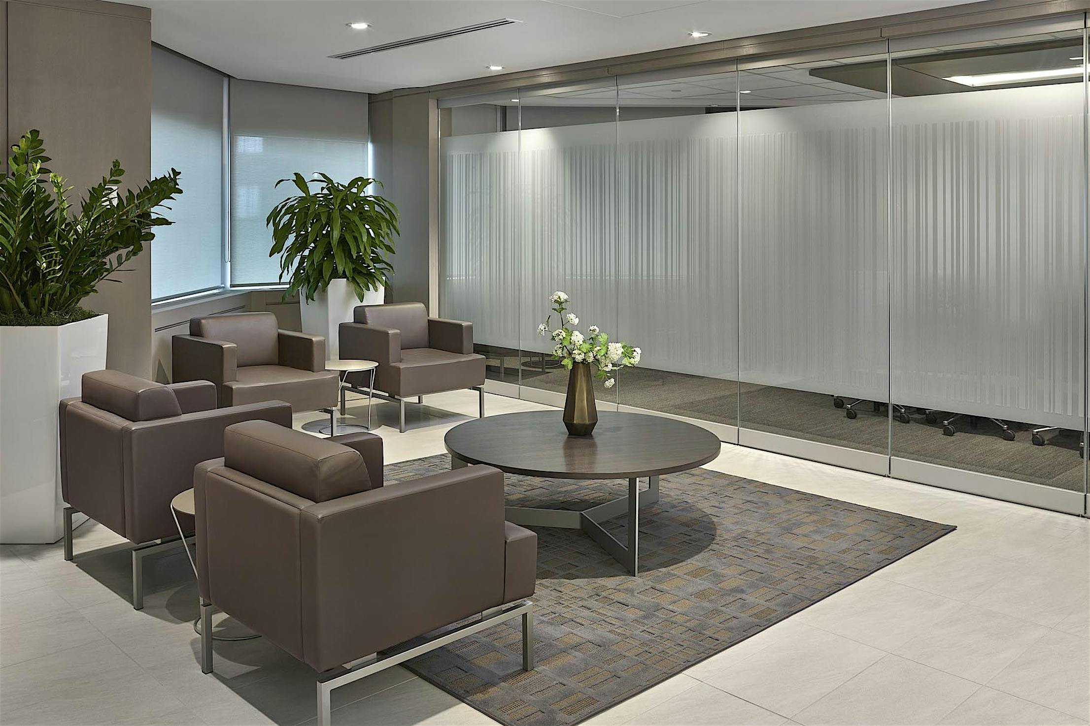Modern office waiting area with brown armchairs and a round coffee table, leading to a conference room with frosted frameless sliding glass doors.