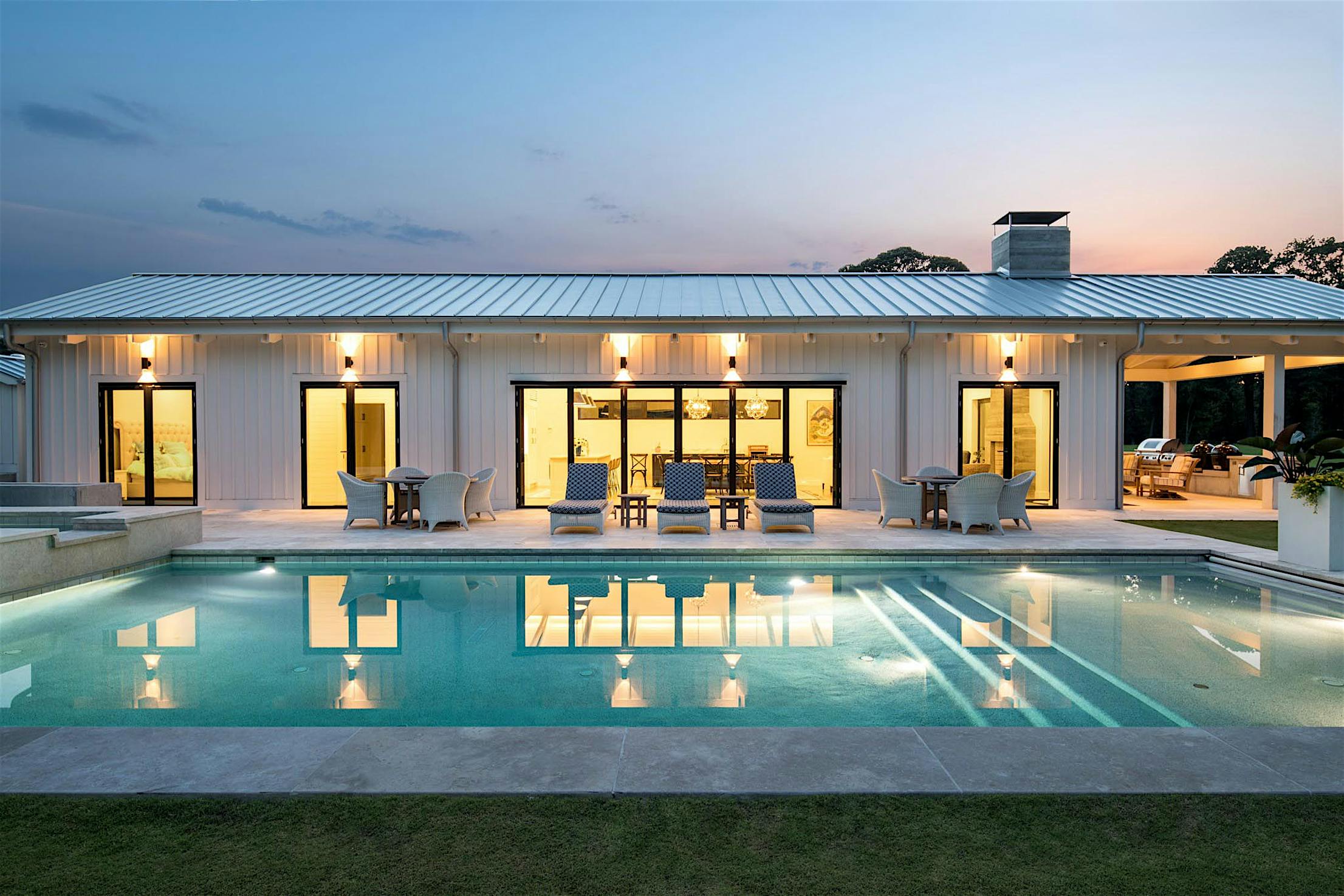 Modern house with bifold glass walls, multiple outdoor seating areas, and an illuminated swimming pool in the foreground at dusk