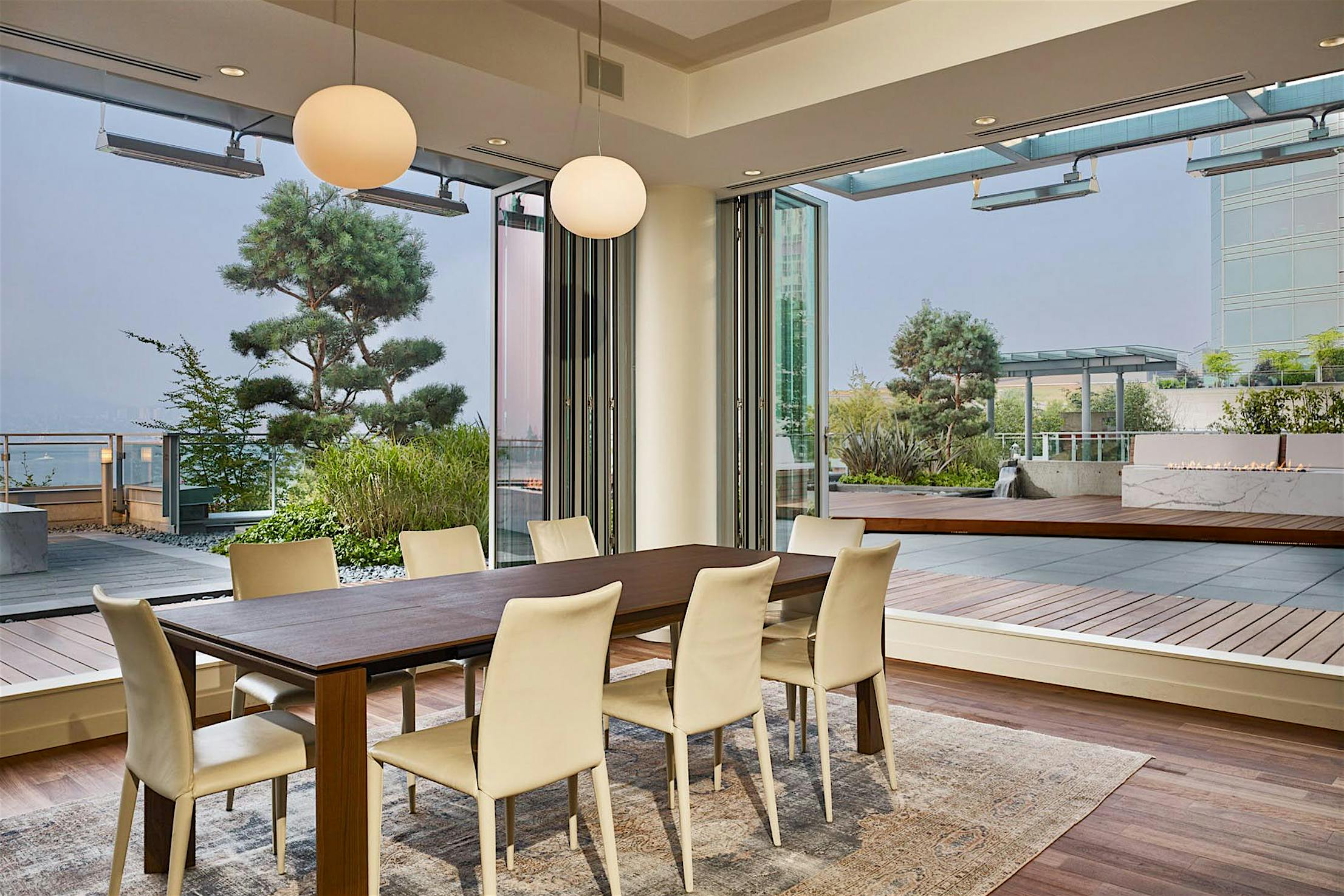 Modern dining area with a wooden table and eight white chairs, featuring accordion glass doors that open to a spacious outdoor patio with greenery.