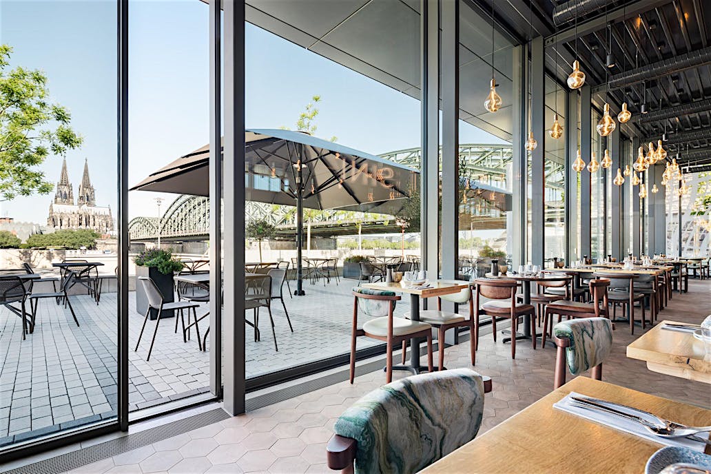 Modern restaurant with floor-to-ceiling glass doors, cityscape views of a cathedral and bridge, and patio seating.