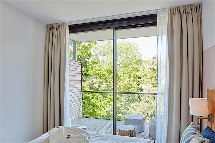 Modern bedroom with double bed, bedside lamp, robes, and floor-to-ceiling windows opening to a balcony with stools and tree views.