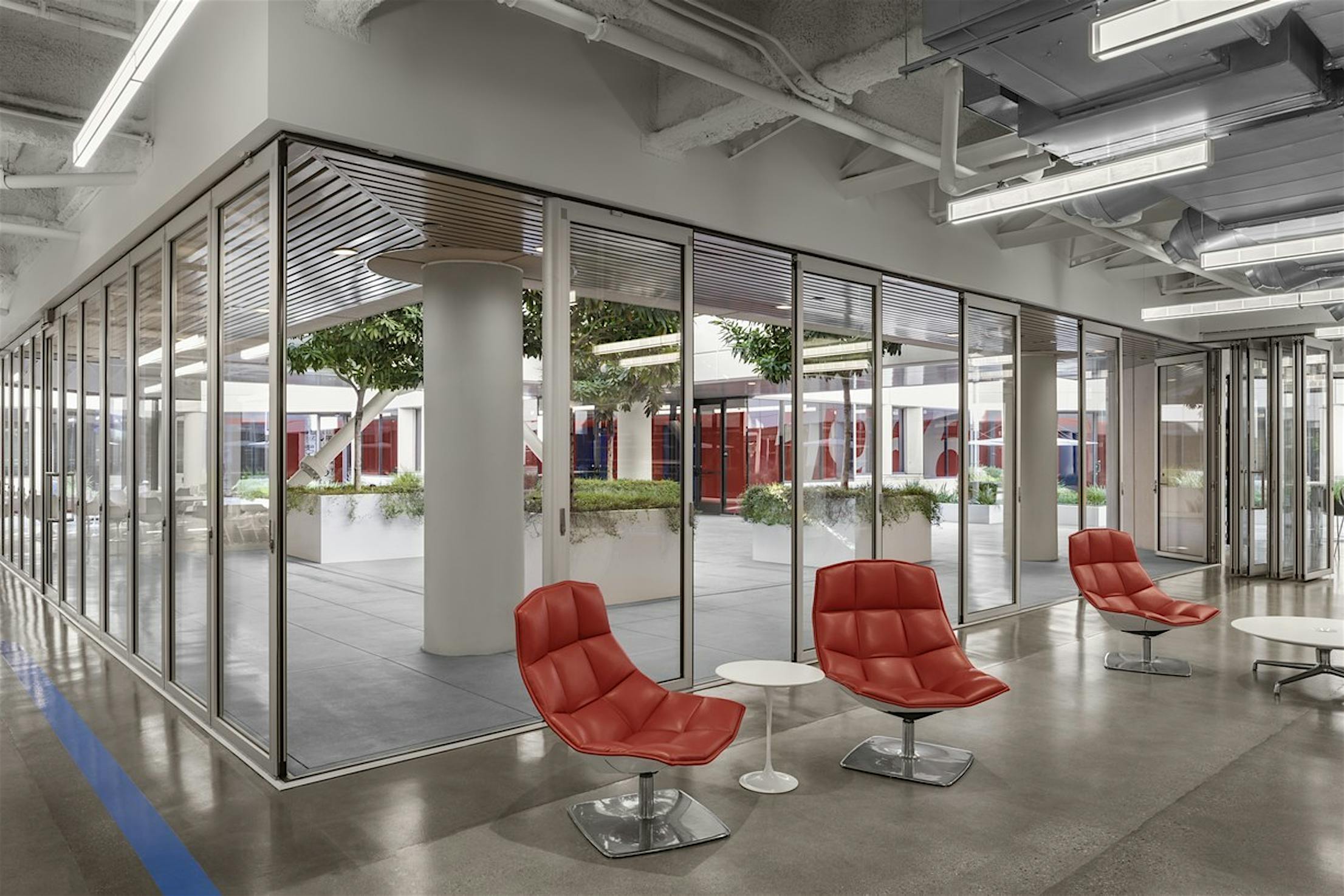 A modern office space with sleek glass office doors, red chairs, white tables, and an indoor garden.