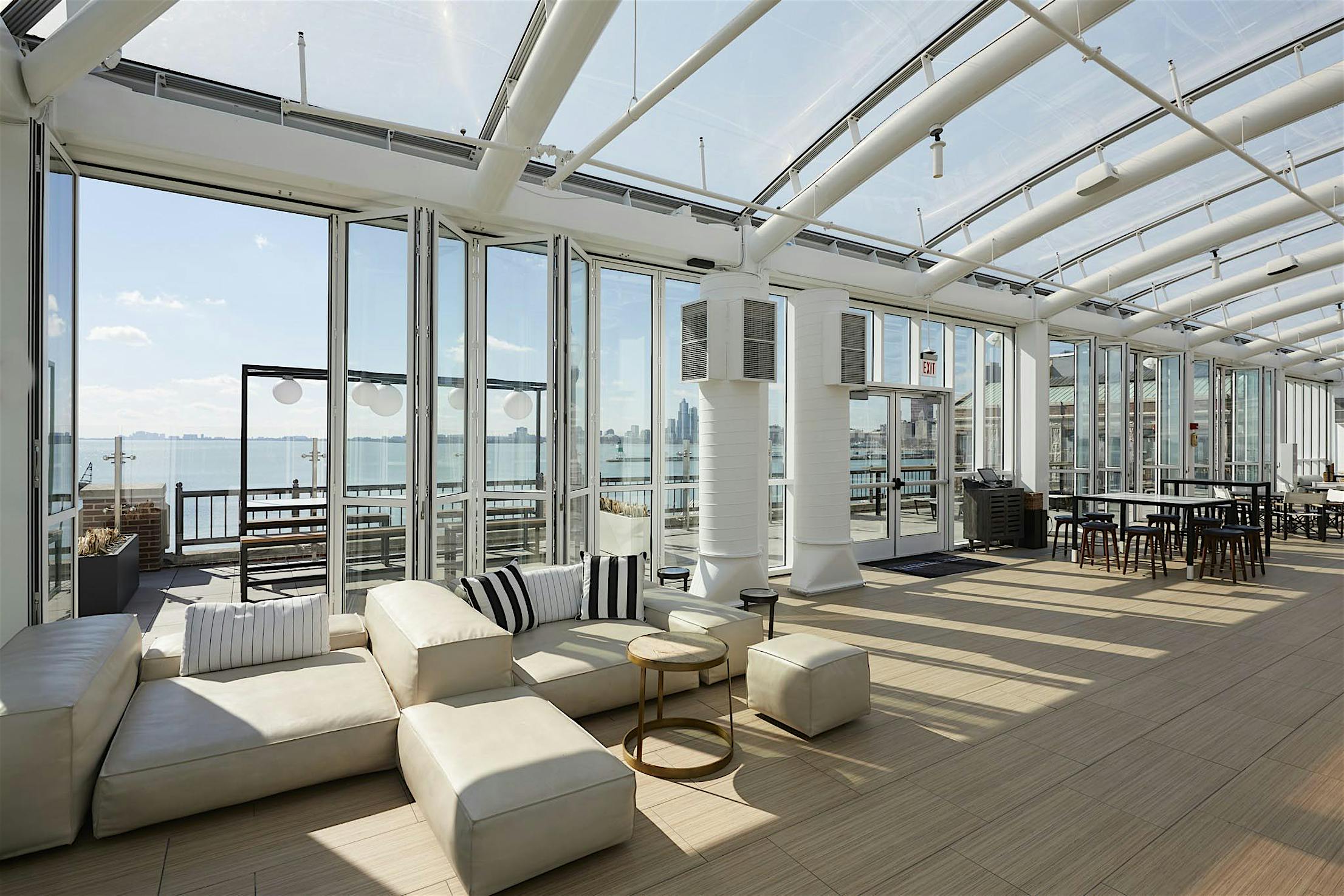 A spacious, modern offshore restaurant with large opening glass walls, light-colored furniture, and wooden flooring. The room opens to a coastal view with seating areas and a clear blue sky visible outside.